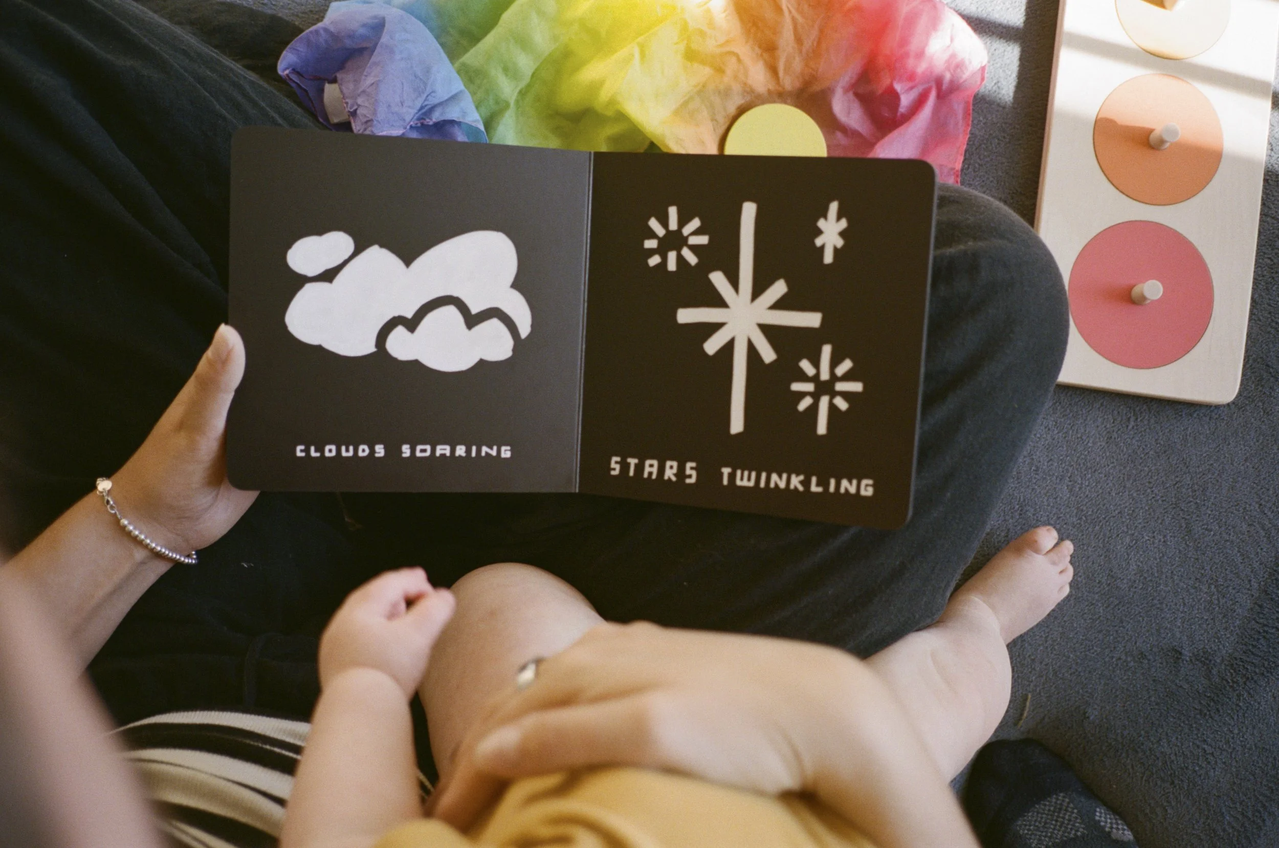 A person sitting on the floor holding an open black card with white illustrations and text that reads "Clouds Soaring," "Stars Twinkling" and various cloud and star symbols. The person has a bracelet on their left wrist, and another person with a bare arm is reaching towards the card. There is a multicolored tissue paper and a wooden toy with colored circular discs in the background.