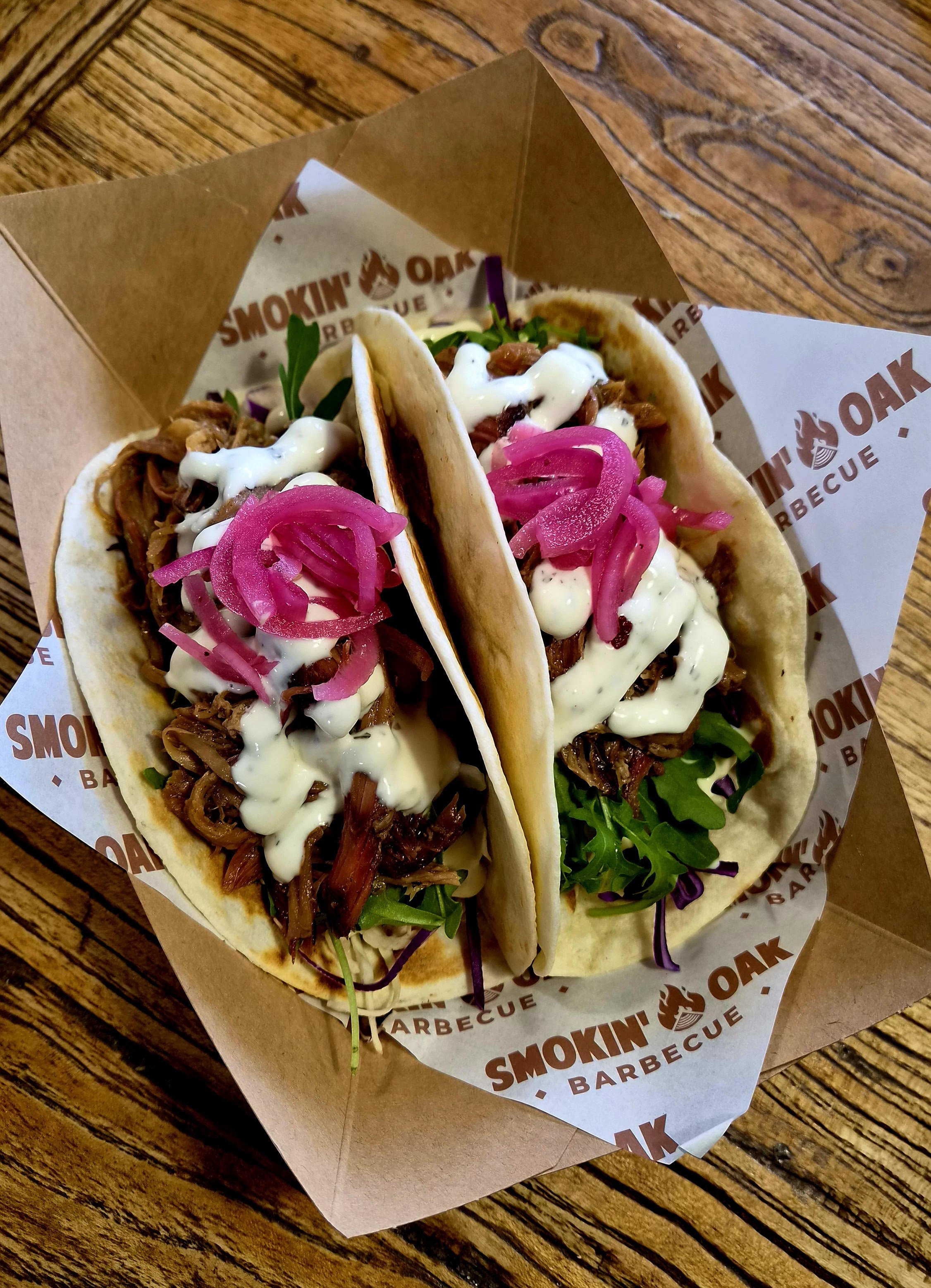 Three tacos filled with shredded meat, pickled onions, and sauce, presented in a paper tray with 'Smokin Oak BBQ' branding, on a wooden surface.