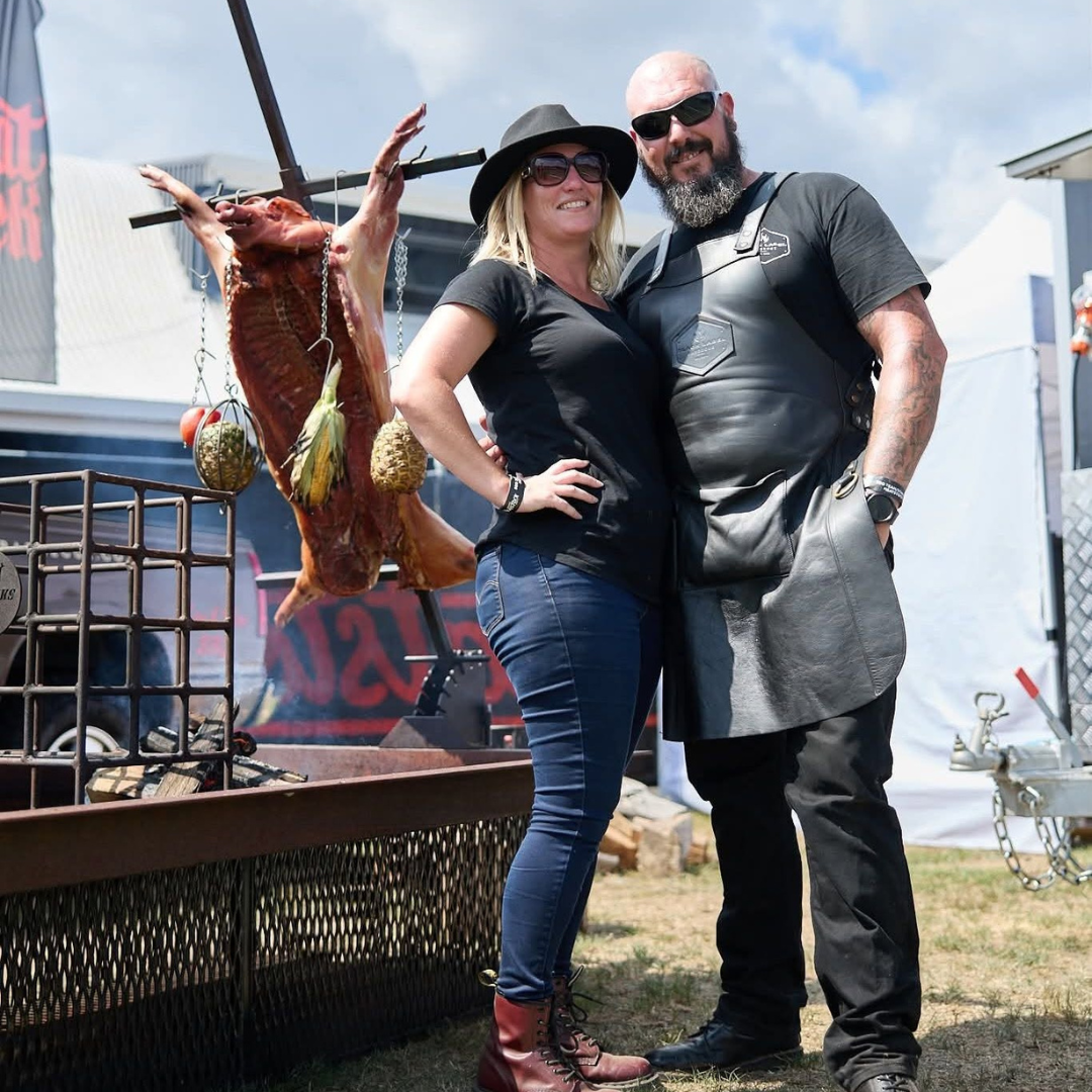 A woman and a man pose together outdoors, with a whole roasted pig hanging behind them, decorated with meat hooks and metal balls, at an outdoor event under a partly cloudy sky.