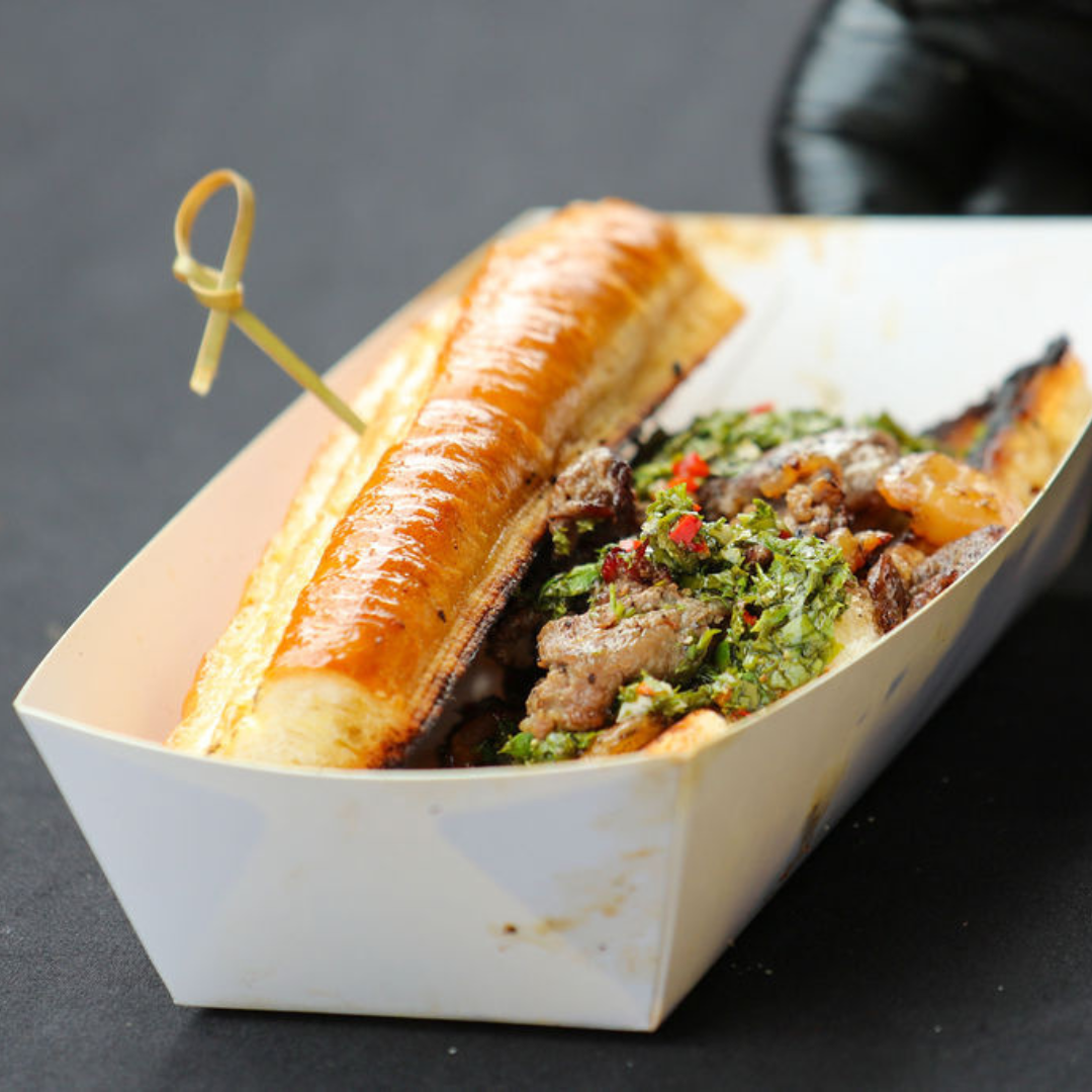 Hot dog with grilled bun and toppings, served in a white paper tray.