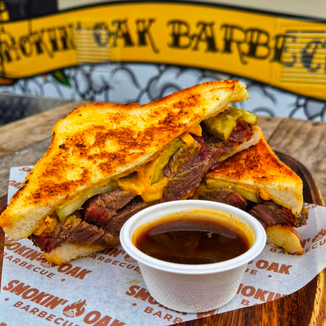A grilled sandwich with beef, cheese, and pickles, served with a side of barbecue sauce on a white paper with 'Smoking Oak Barbecue' printed on it.