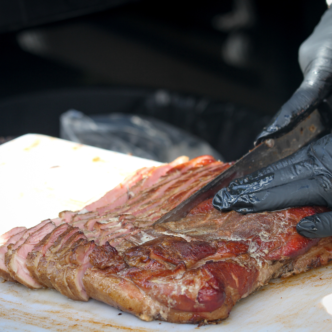 A person wearing black gloves slicing cooked bacon on a white cutting board.