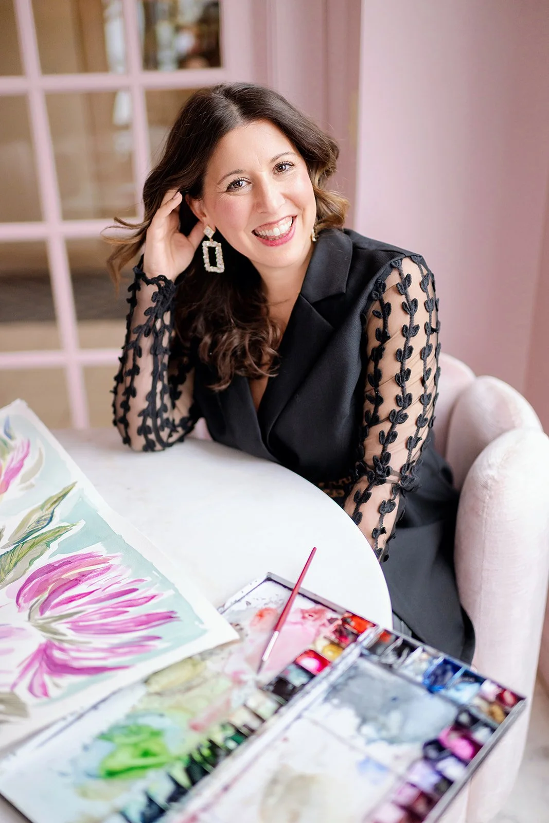 Local artist and founder Giovanna Chaisson wearing a black top sits smiling at a white table with her painting in front of her