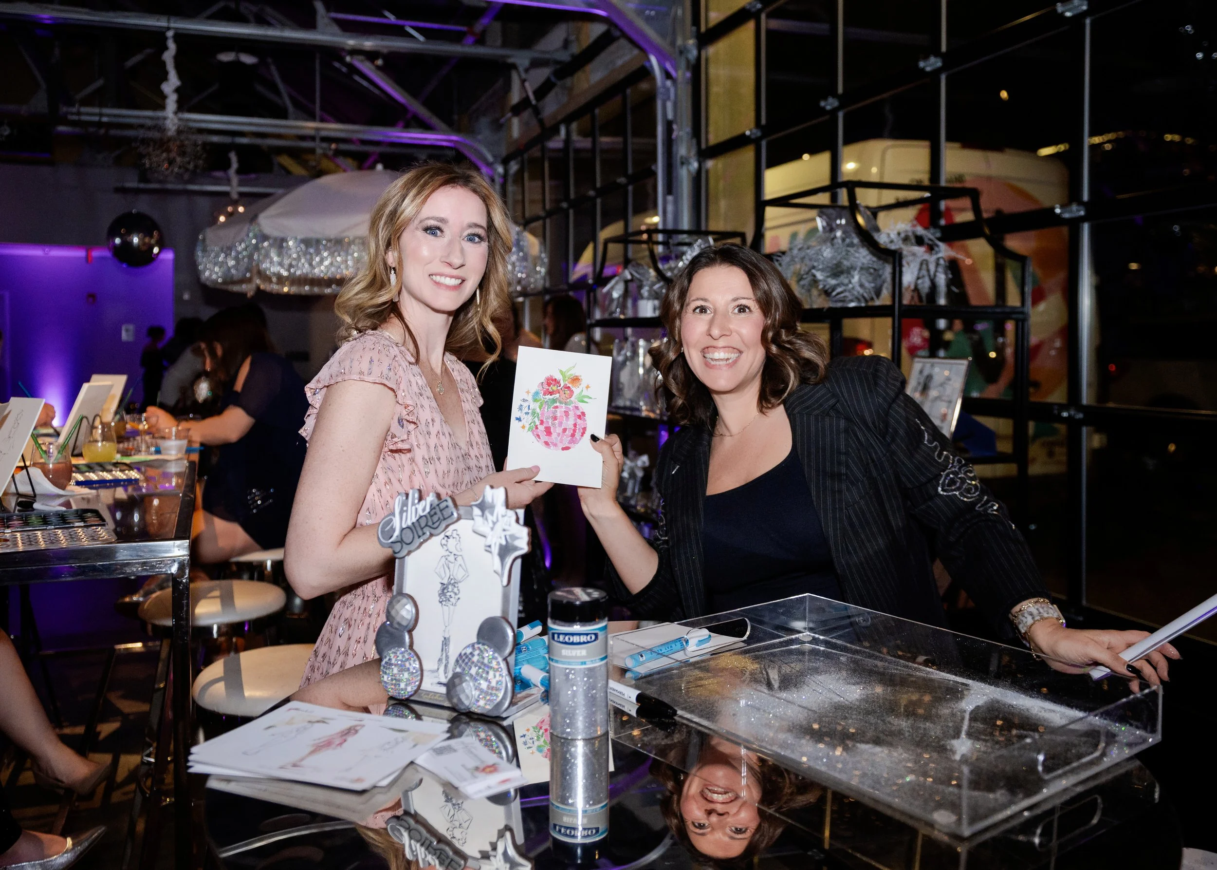 Two women smile at the camera while holding up their artwork at a silver themed event