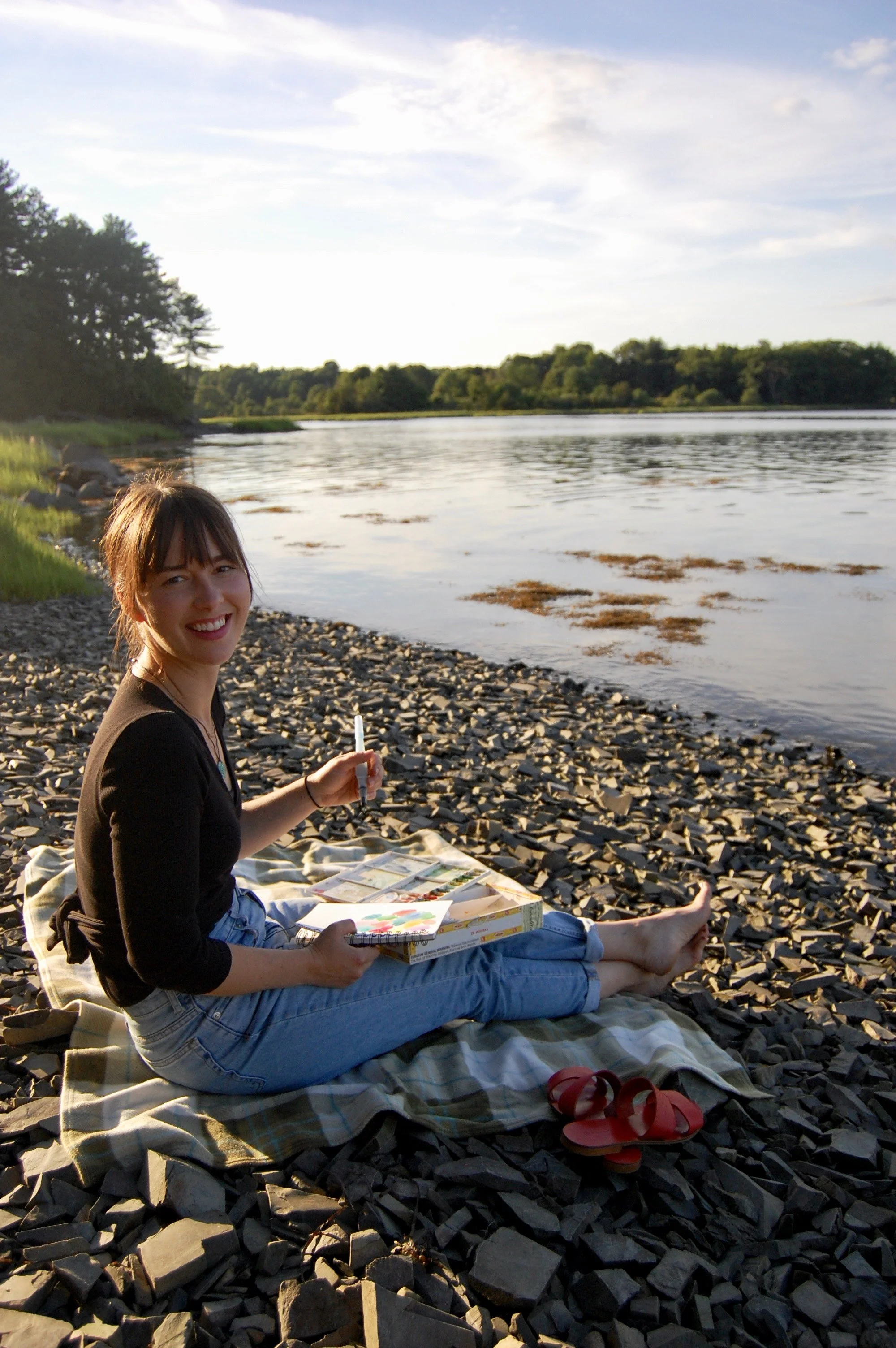 Woman smiles while painting on the edge of a rocky beach
