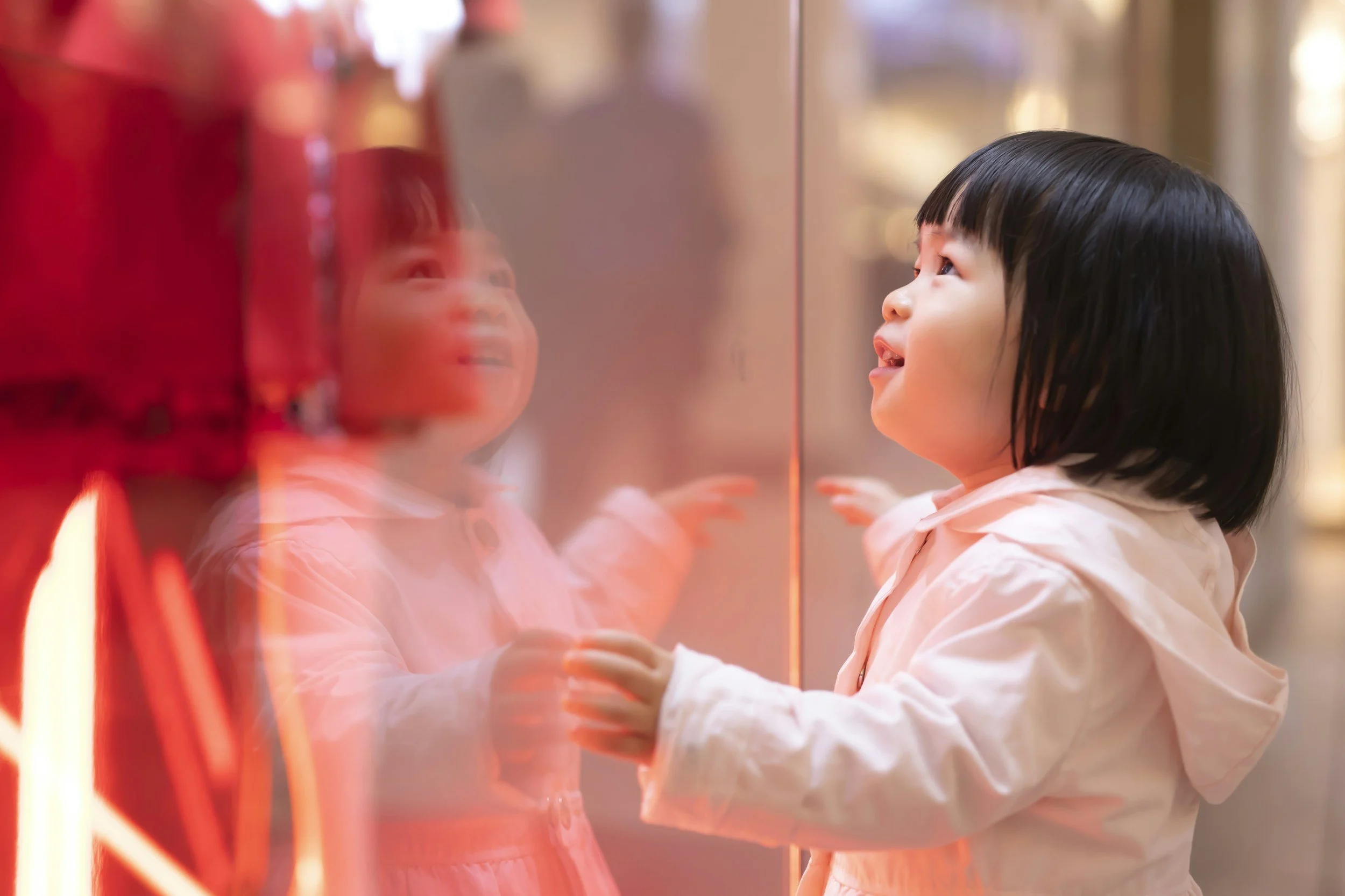 Young girl with short black hair looking and smiling at her reflection in a glass display case.