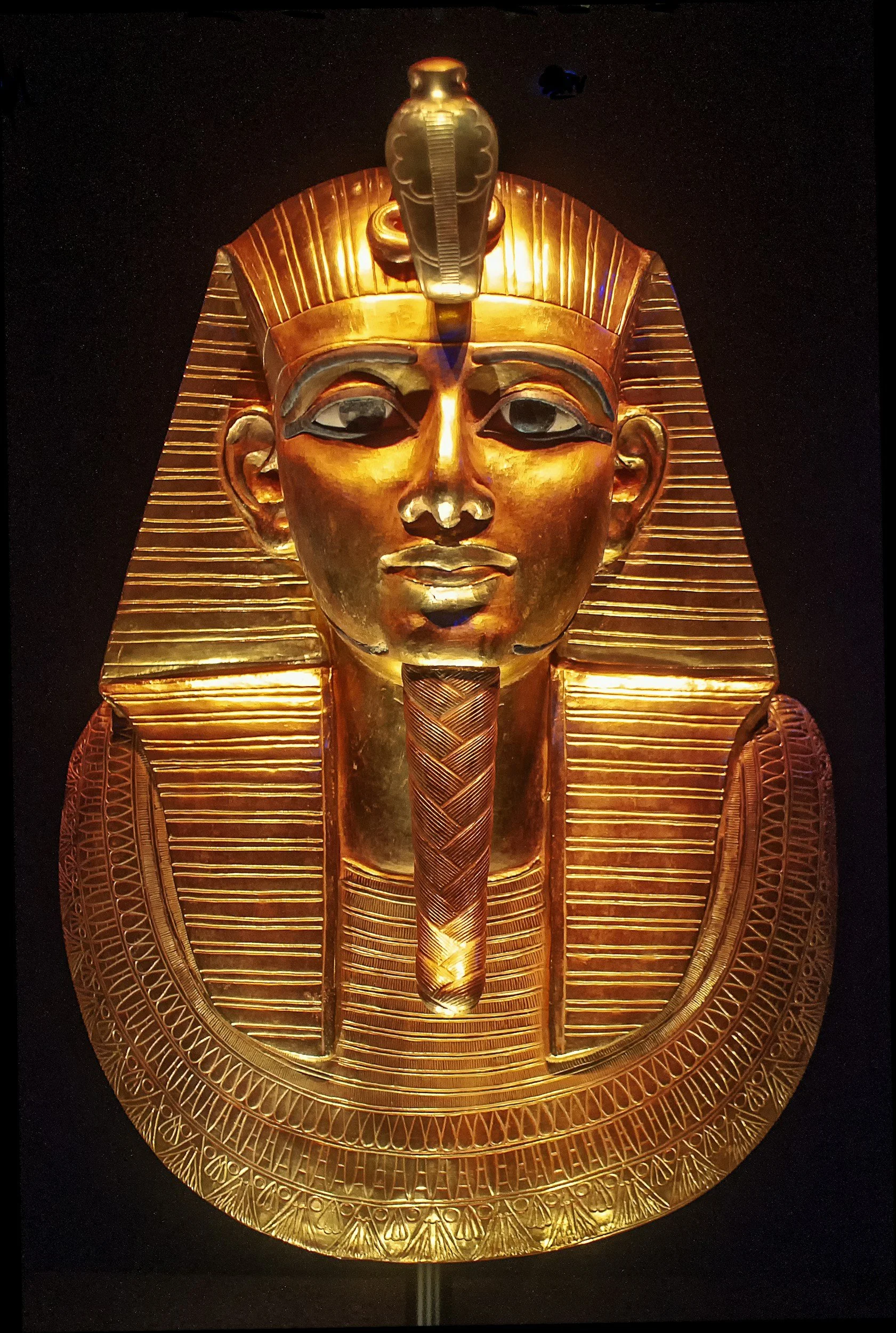 Gold funeral mask of an ancient Egyptian pharaoh, featuring a striped nemes headdress, a snake-shaped uraeus on the forehead, and detailed carvings.