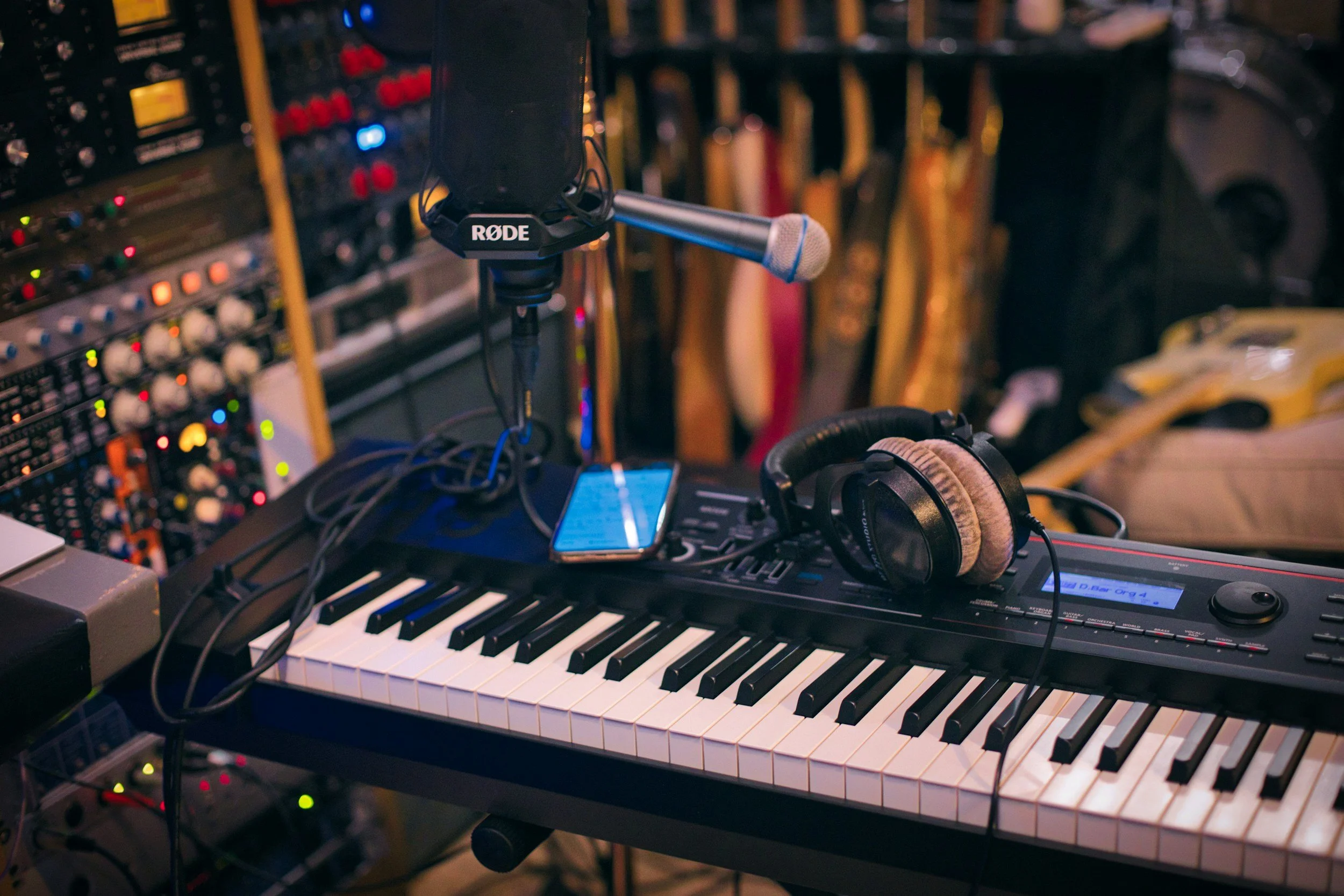 A keyboard with headphones resting on it, a microphone attached to a stand, and a smartphone placed on the keyboard, with audio equipment and guitars in the background.