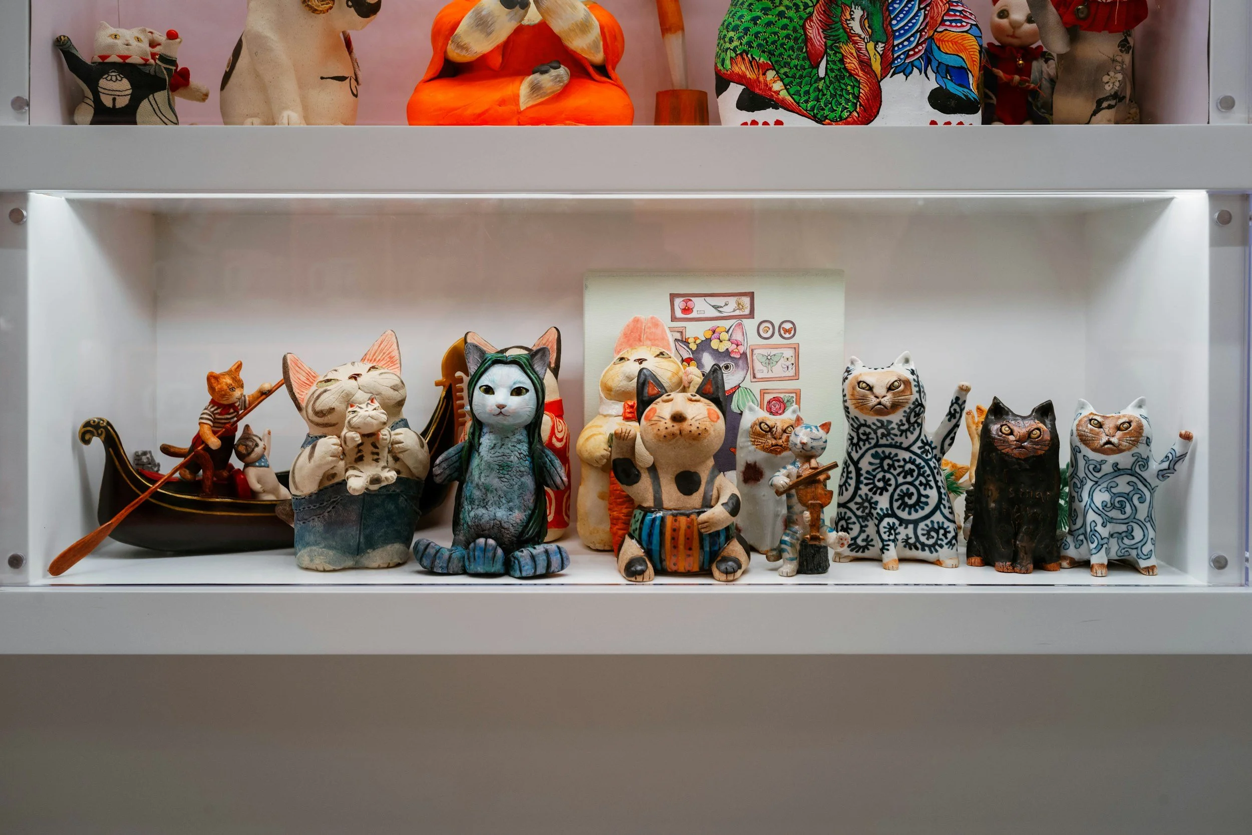 Collection of cat-themed figurines and artwork displayed on a white shelf, featuring various cats in different styles and poses, including traditional, whimsical, and artistic designs.