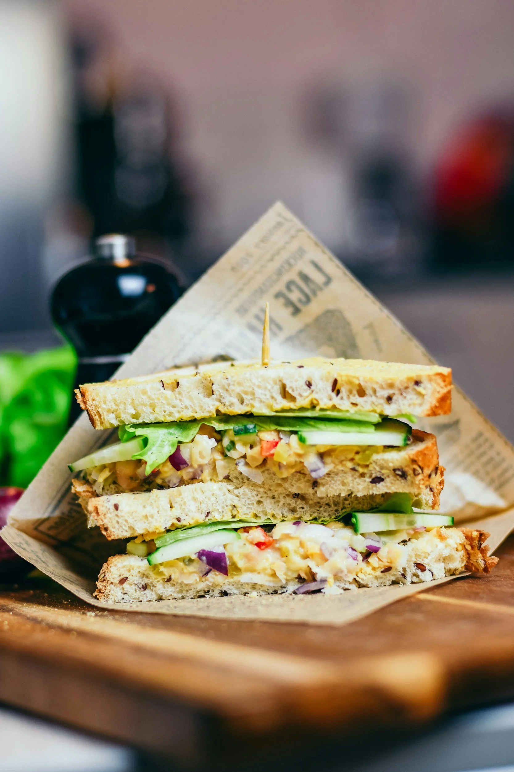 A triple-layered sandwich with lettuce, cucumber, and a spread, served on whole grain bread, with a toothpick holding it together. The sandwich is on a wooden surface with a slice of bread on top.