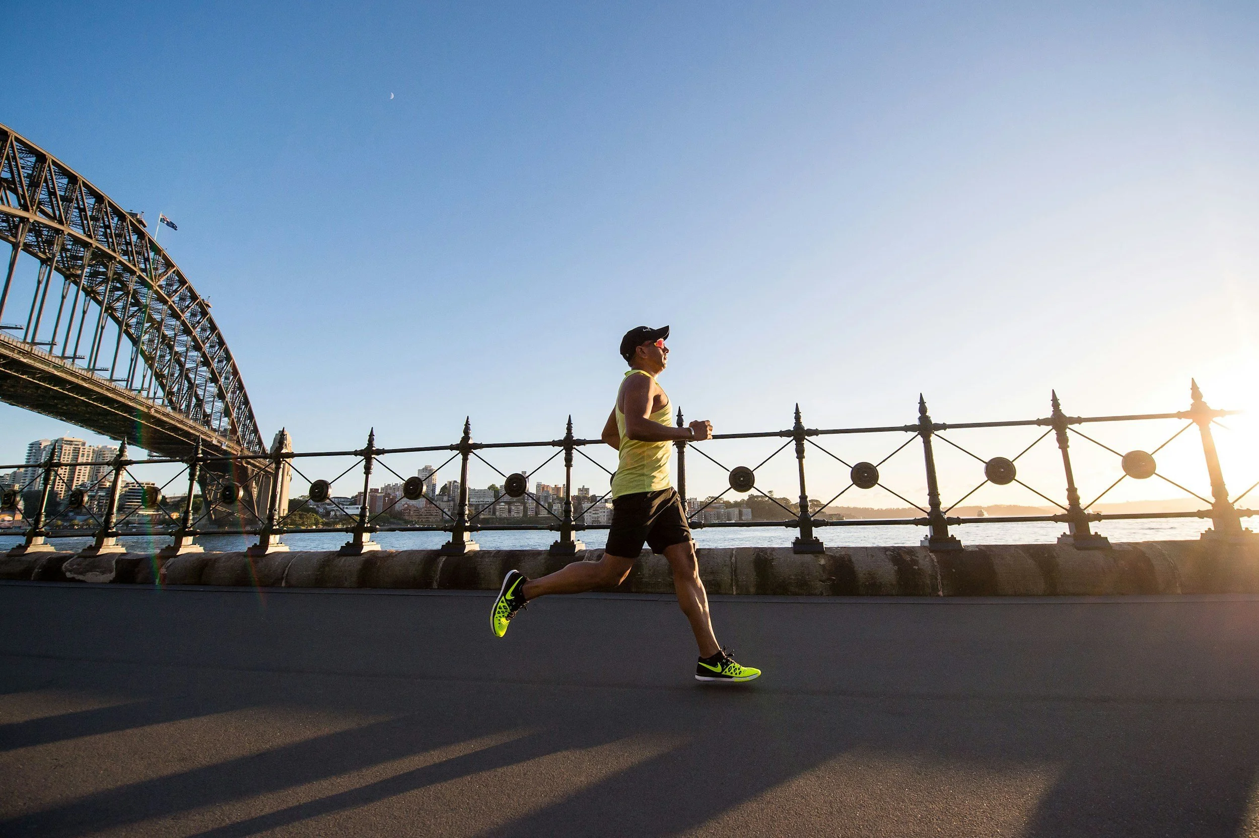 A man running along a riverfront path with a bridge and city skyline in the background during sunset.