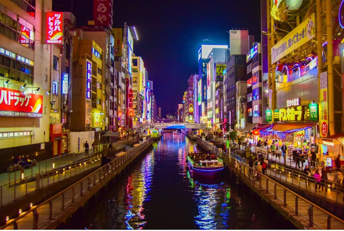 A boat on a city river at night, buildings that line the river have bright, neon lights