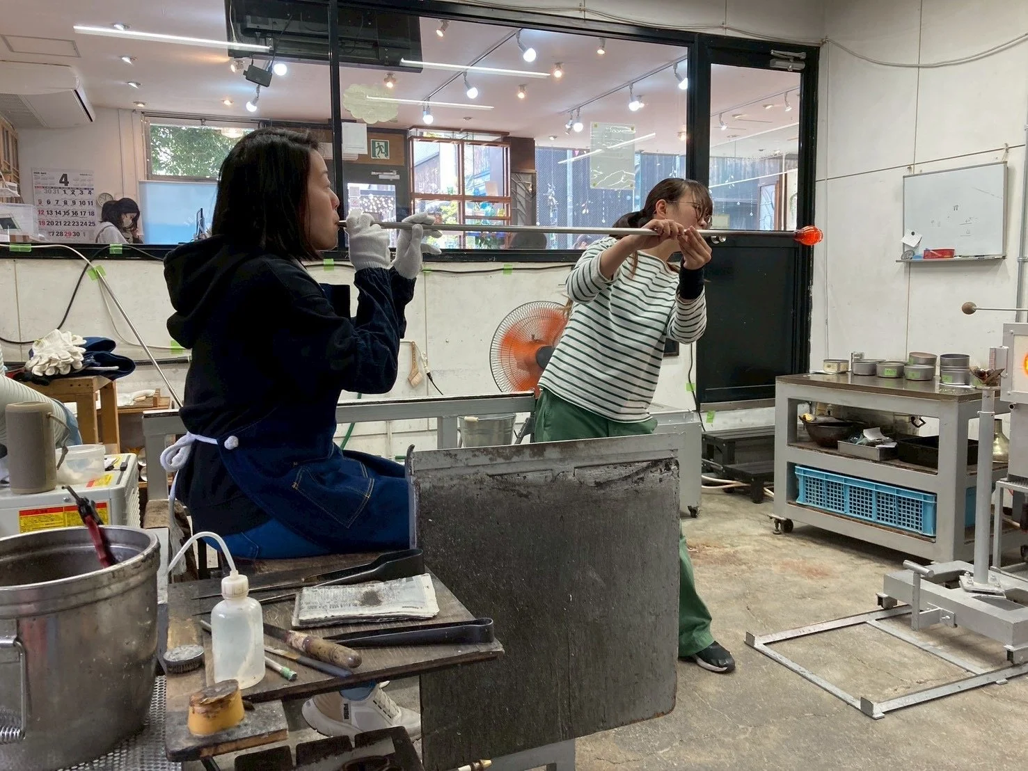 Two women are practicing glassblowing in a studio. One woman is blowing into a pipe while the other assists, holding the pipe steady. Various tools and equipment are visible on the worktable and in the background.
