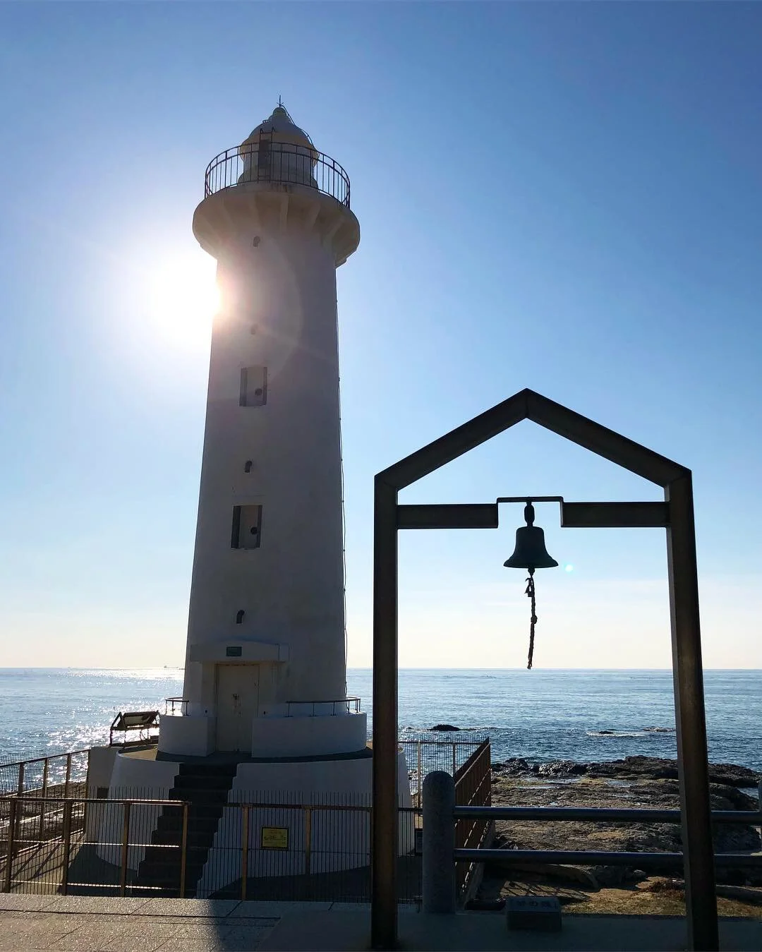 A lighthouse tower backlit by the sun, next to a bell mounted inside an arch, on the rocky shore of the sea.