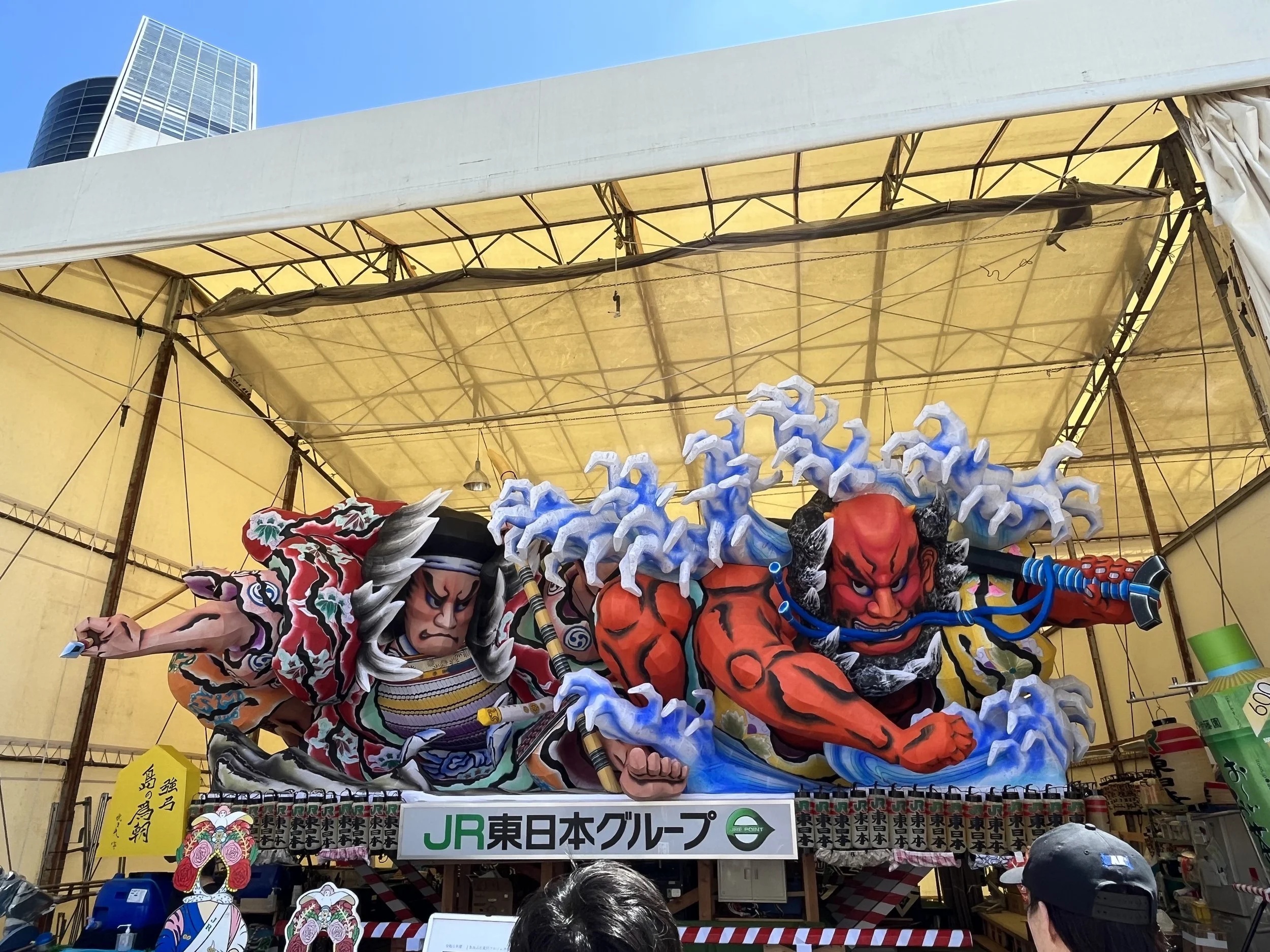 Colorful traditional Japanese festival float featuring fierce warriors with dynamic poses, vibrant costumes, and exaggerated facial expressions, under a yellow tent with modern buildings in the background.