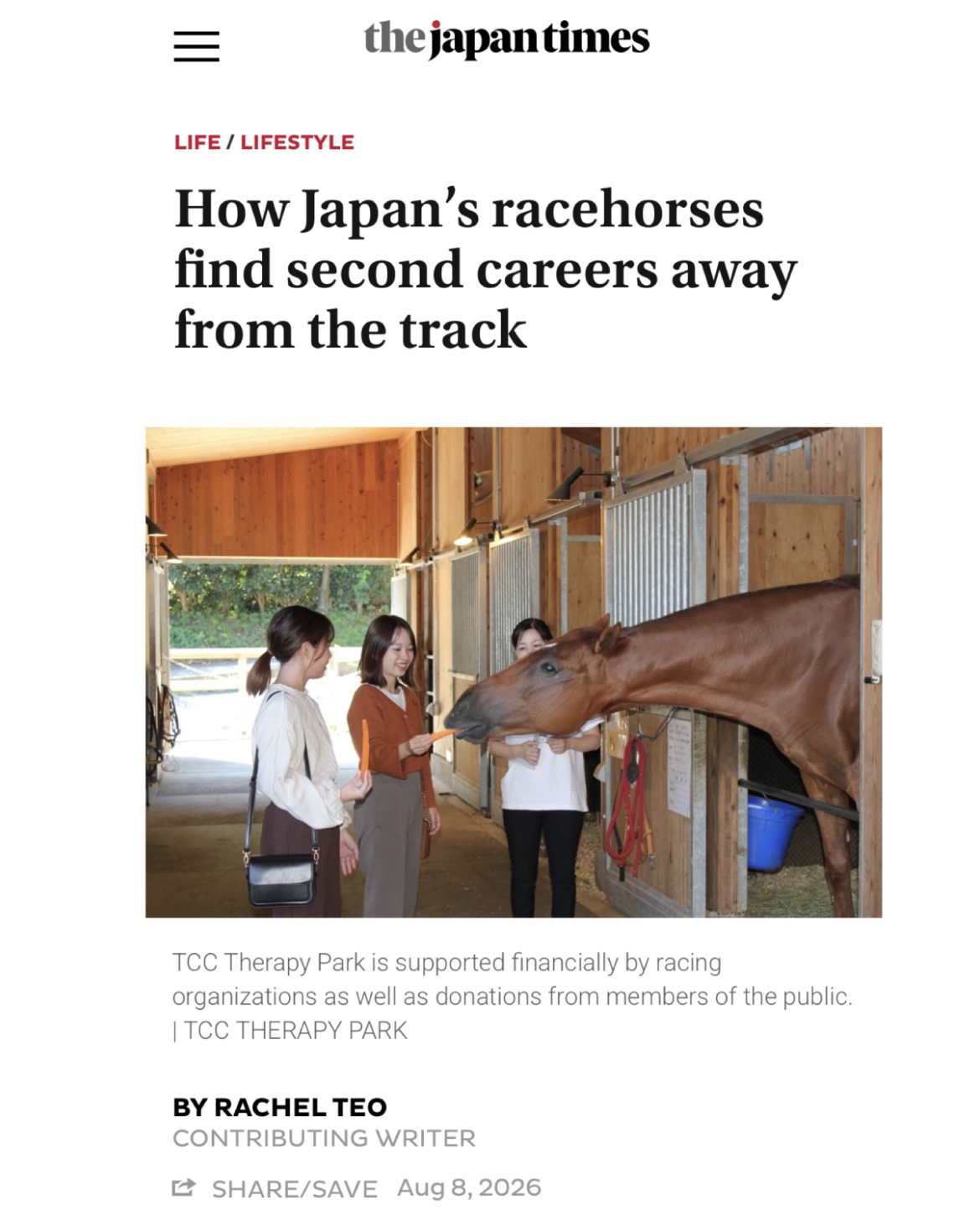 Three women inside a stable are feeding a brown horse a carrot. The stable has wooden walls and stalls.