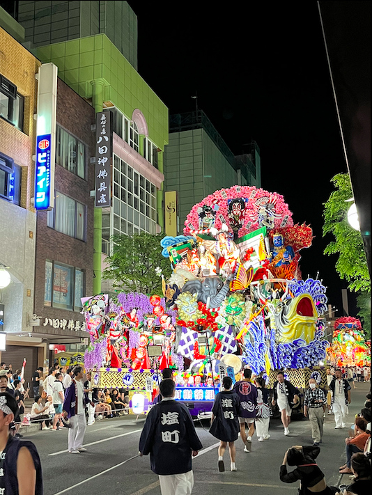 A three-meter tall, ornately decorated, wooden festival float unfolding from its trailer to be about five-meters wide. The float depicts a variety of scenes from Japanese folklore and has demons, gods, an elephant, and dancers dressed in kimonos. 