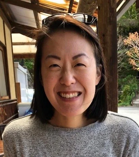 A woman smiling at an outdoor dining area with wooden beams and greenery in the background.