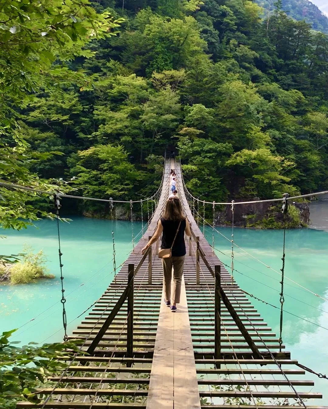 A woman is crossing a wooden and steel suspension bridge over turquoise-coloured water