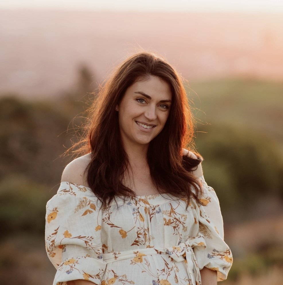 A woman with long brown hair smiling outdoors during sunset, wearing a white off-shoulder floral dress.