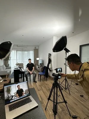 Person filming another person sitting in a living room for a video recording. Lights and camera equipment set up.