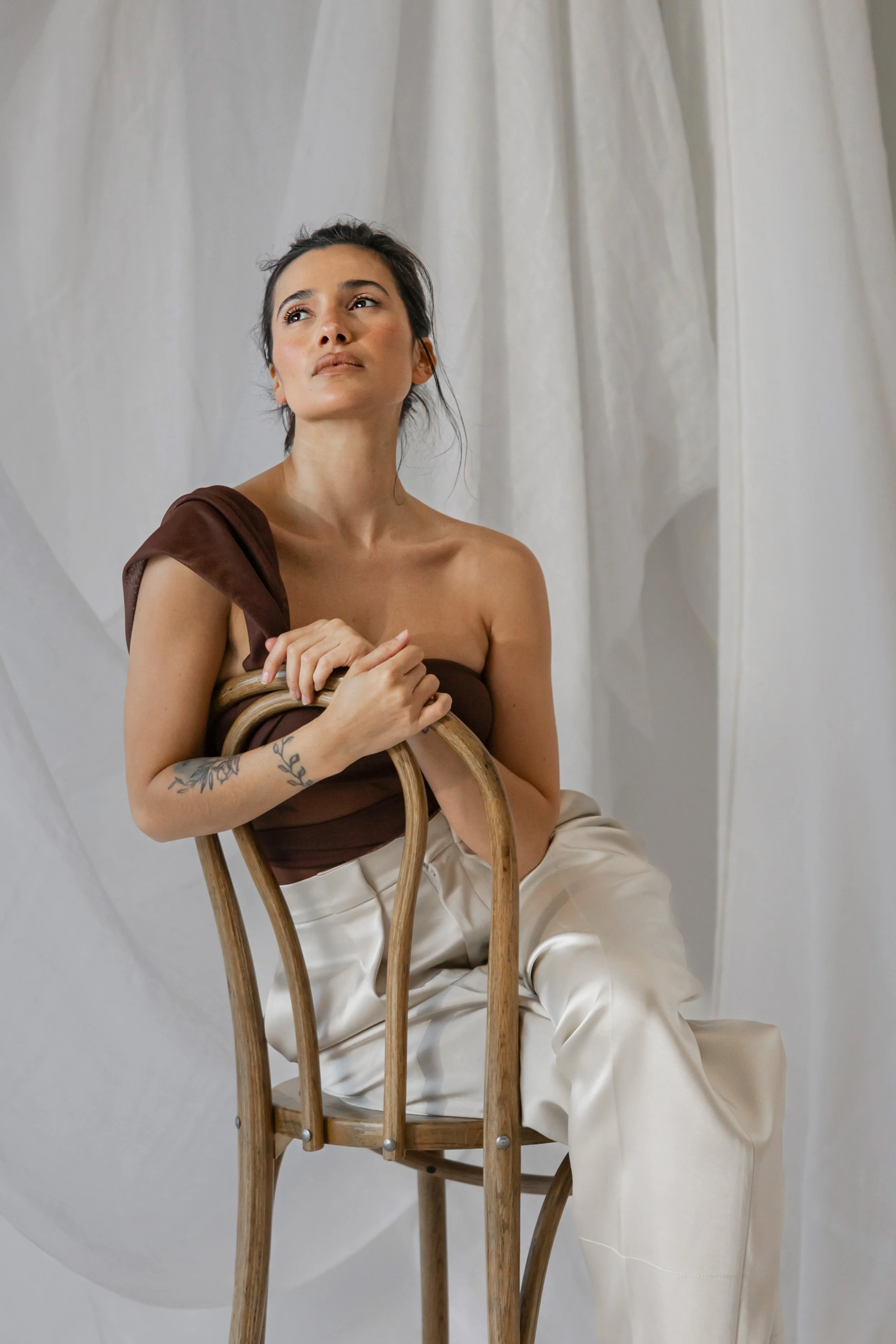 A woman with dark hair and tattoos on her arm, wearing a brown top and cream-colored pants, sitting on a wooden chair against a backdrop of white drapes, looking thoughtfully into the distance.
