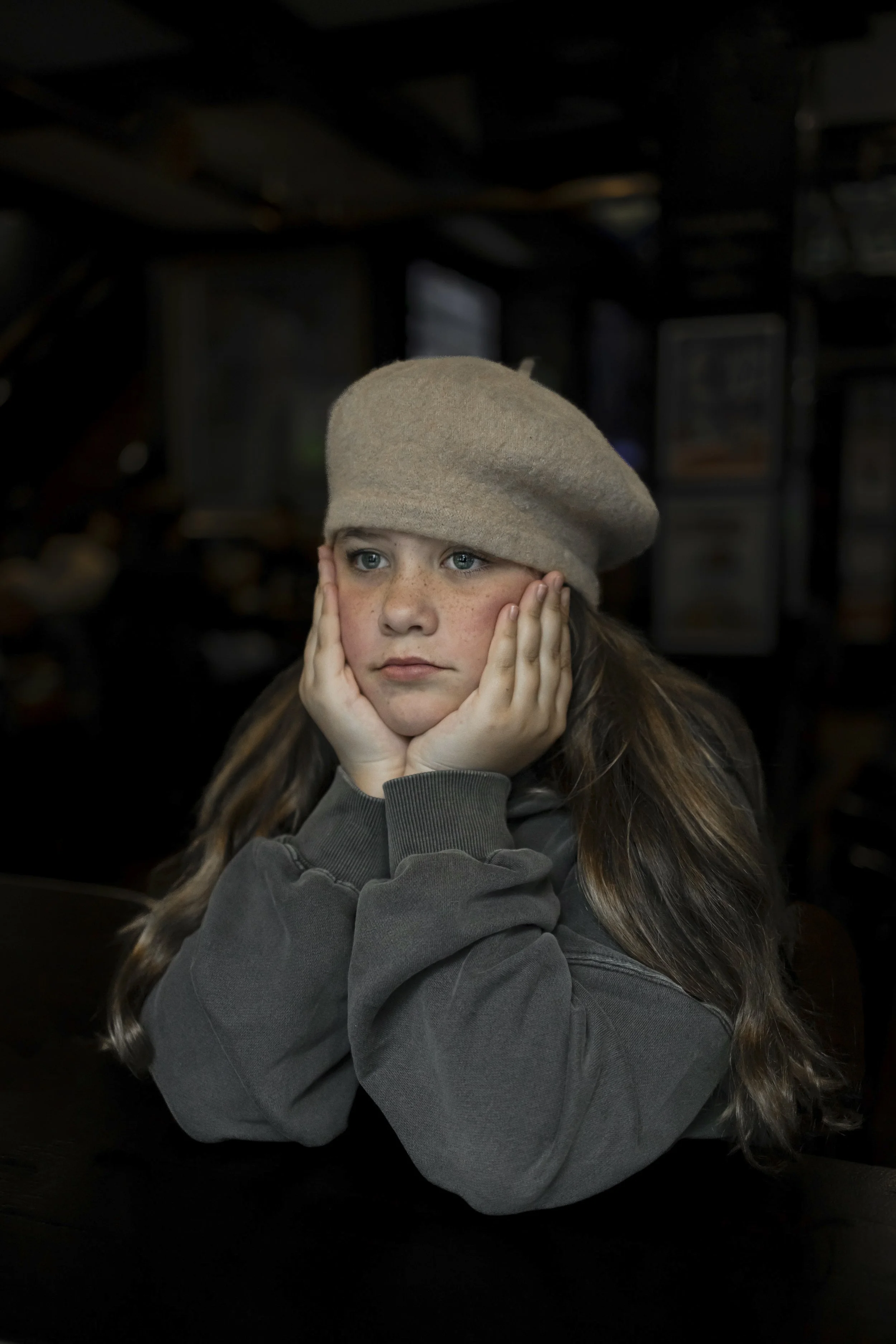 A young girl with blue eyes and freckles resting her face in her hands, wearing a beige beret and a dark gray sweatshirt, sitting in a dimly lit room.