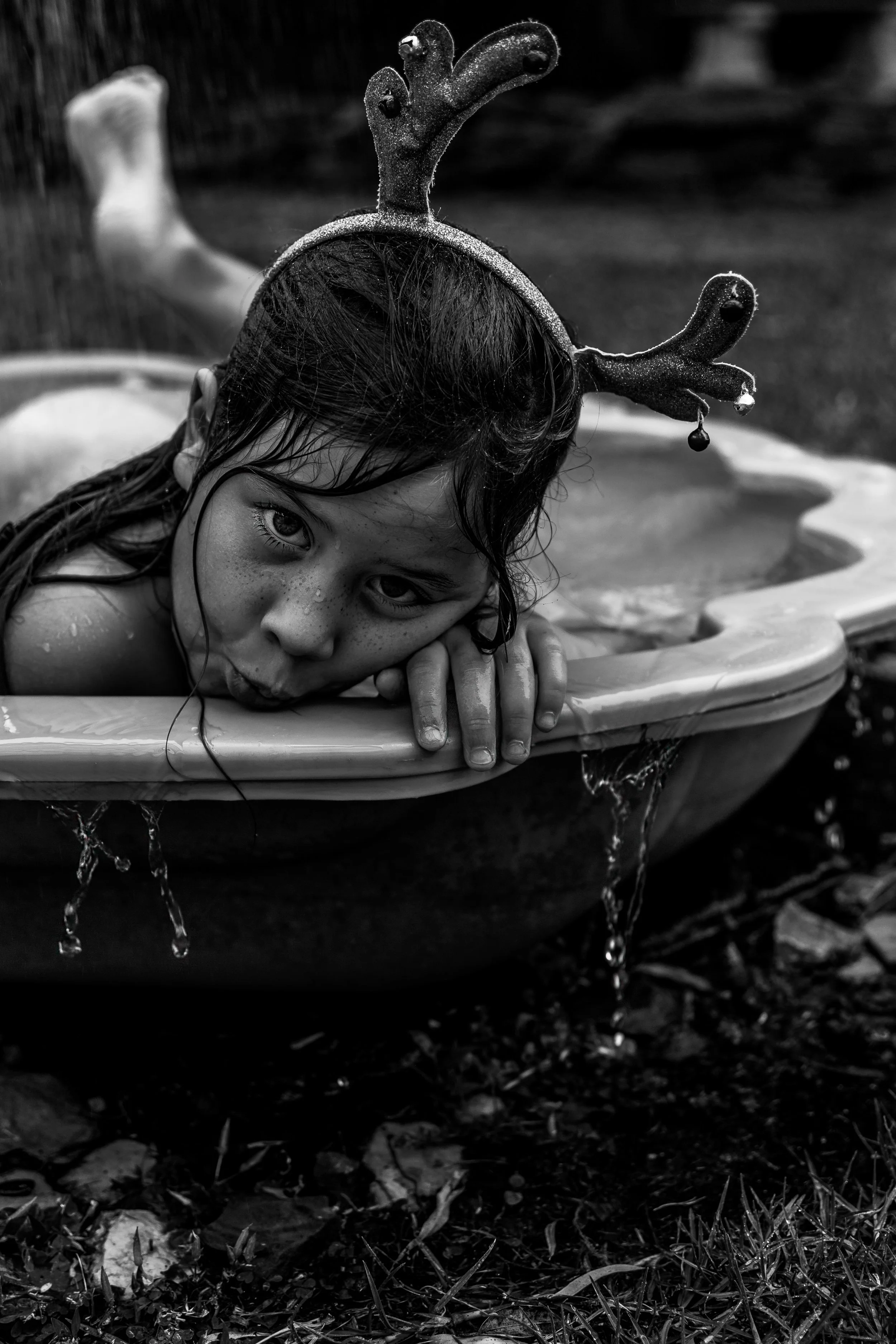 A young girl with wet hair resting her head on the edge of a small plastic kiddie pool, wearing reindeer antler headband, outdoors on grass and leaves, black and white photo.