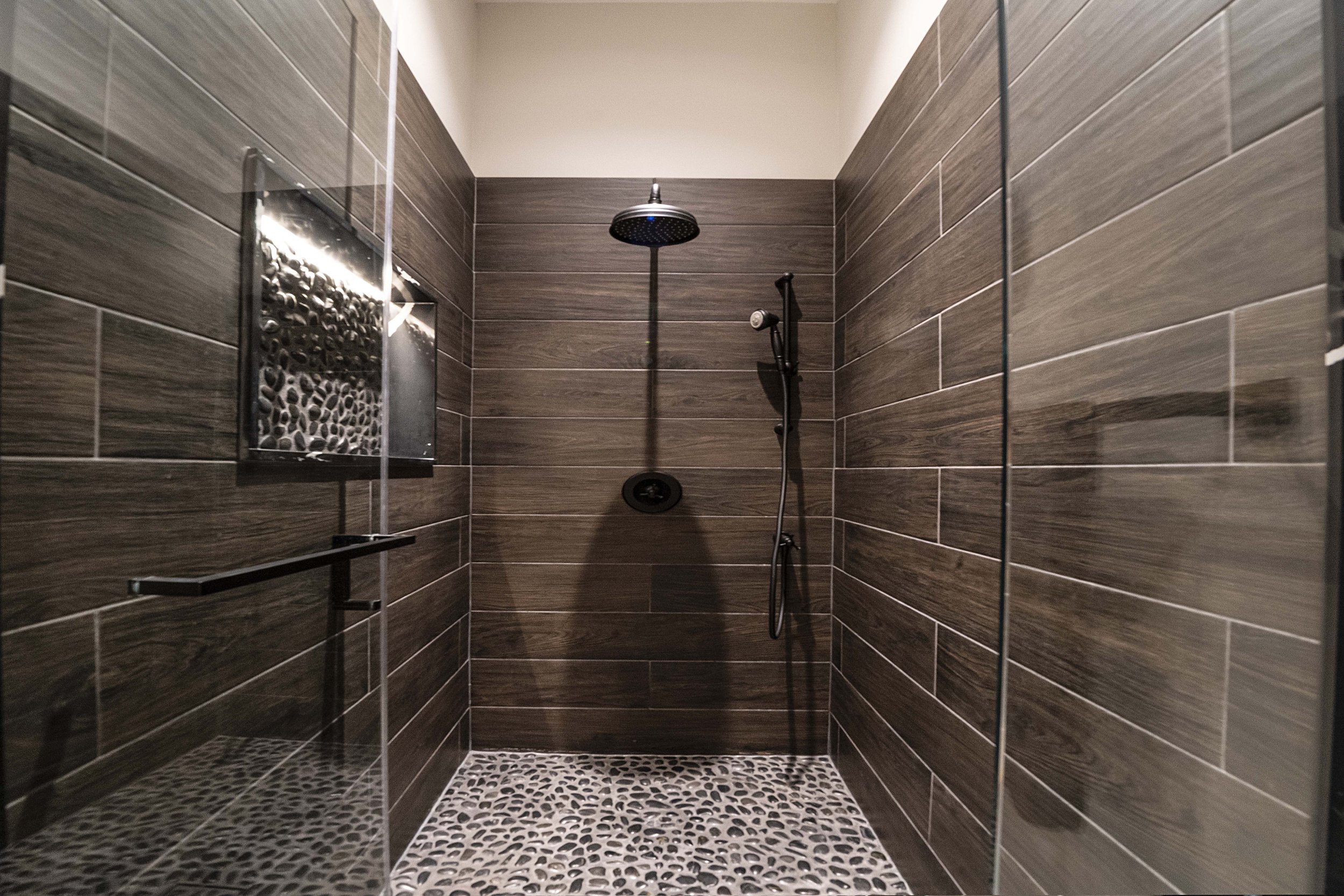 Modern shower with dark wood-like tile walls, pebble floor, rainfall showerhead, and handheld shower wand.