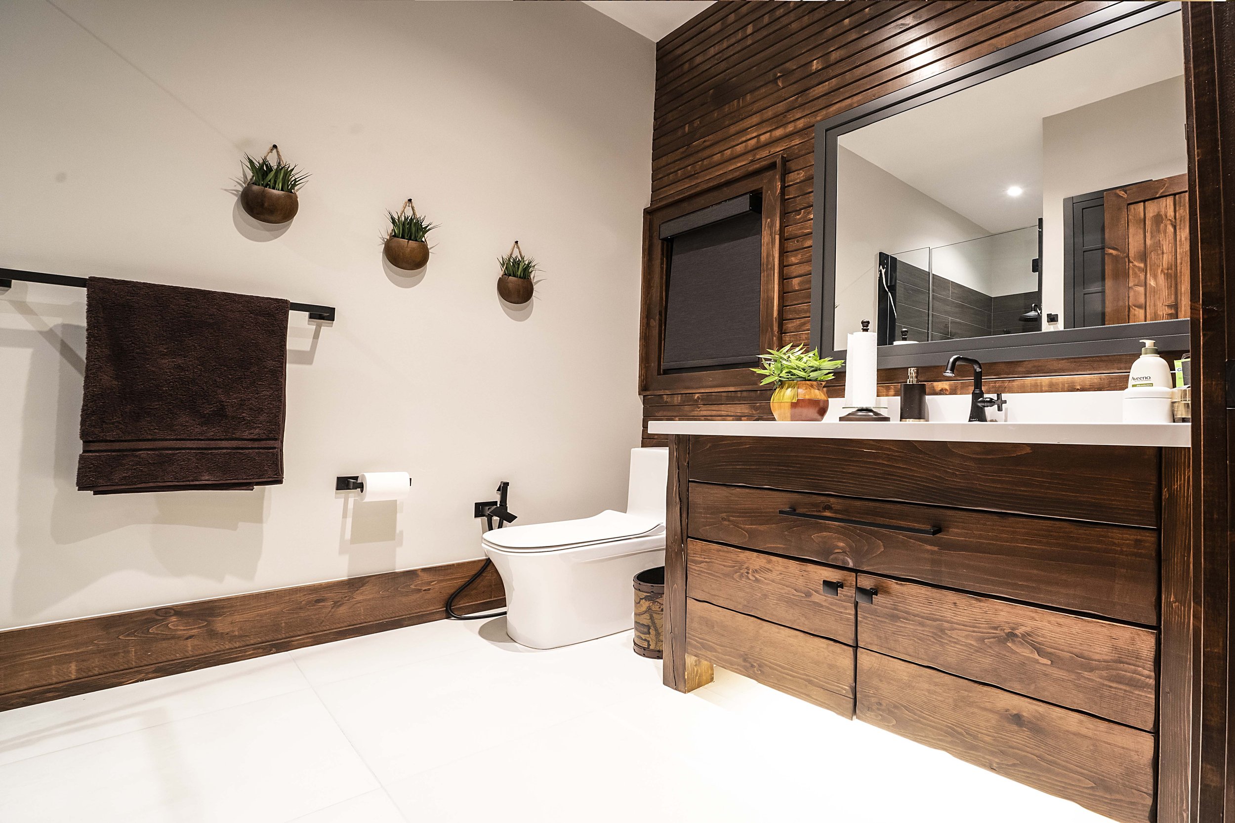 Modern bathroom with wooden accents, featuring a toilet, sink with a wooden vanity, wall-mounted faucet, dark brown towel, wall-mounted planters, and a large mirror.