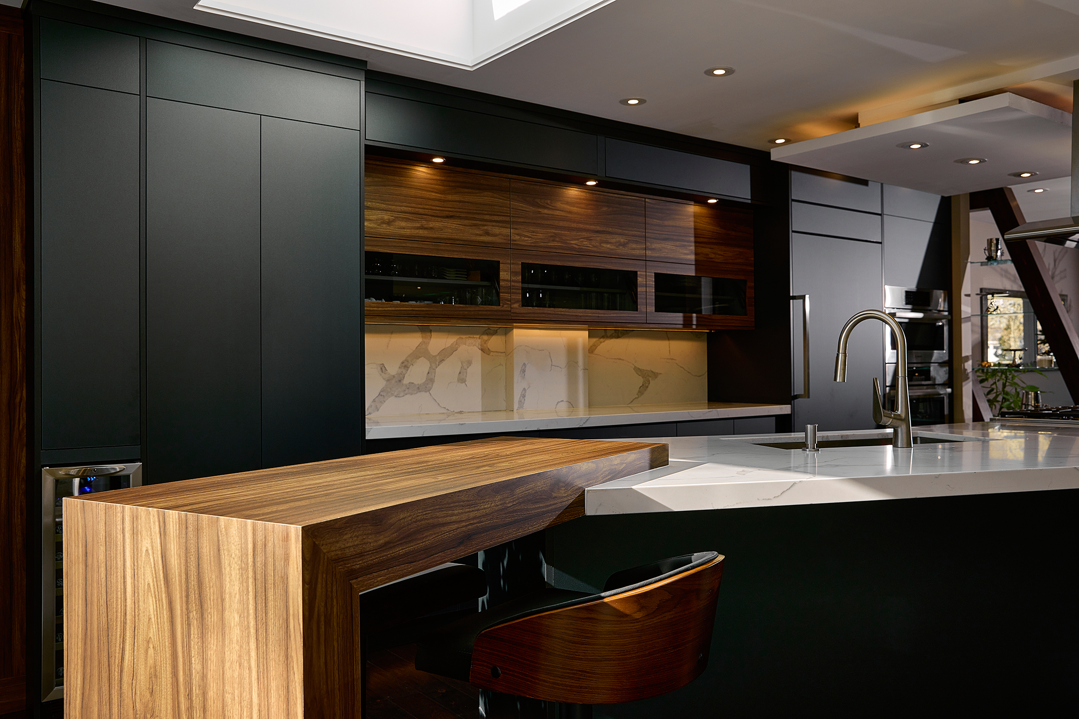 Modern kitchen with black cabinets, wood-paneled accents, marble countertops, and stainless steel appliances. Features include a sleek faucet, under-cabinet lighting, and a geometric bar counter with a contemporary chair.