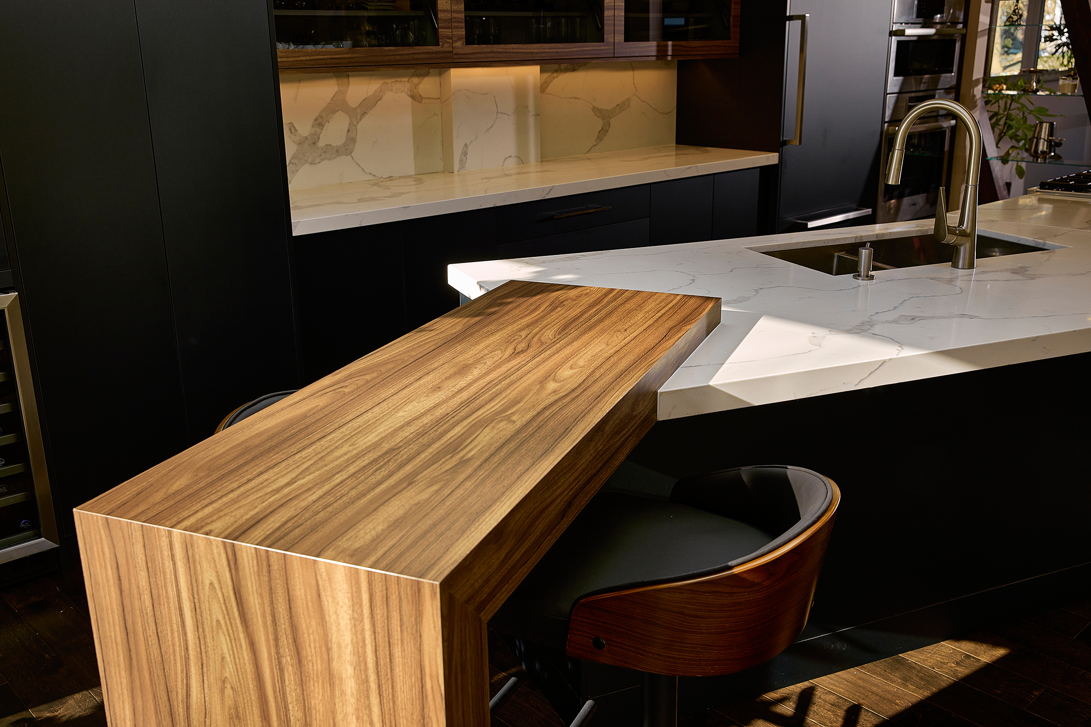 Modern kitchen interior with marble countertops, a wooden breakfast bar, and a stainless steel sink. Dark cabinetry and wall panels enhance the contemporary design.
