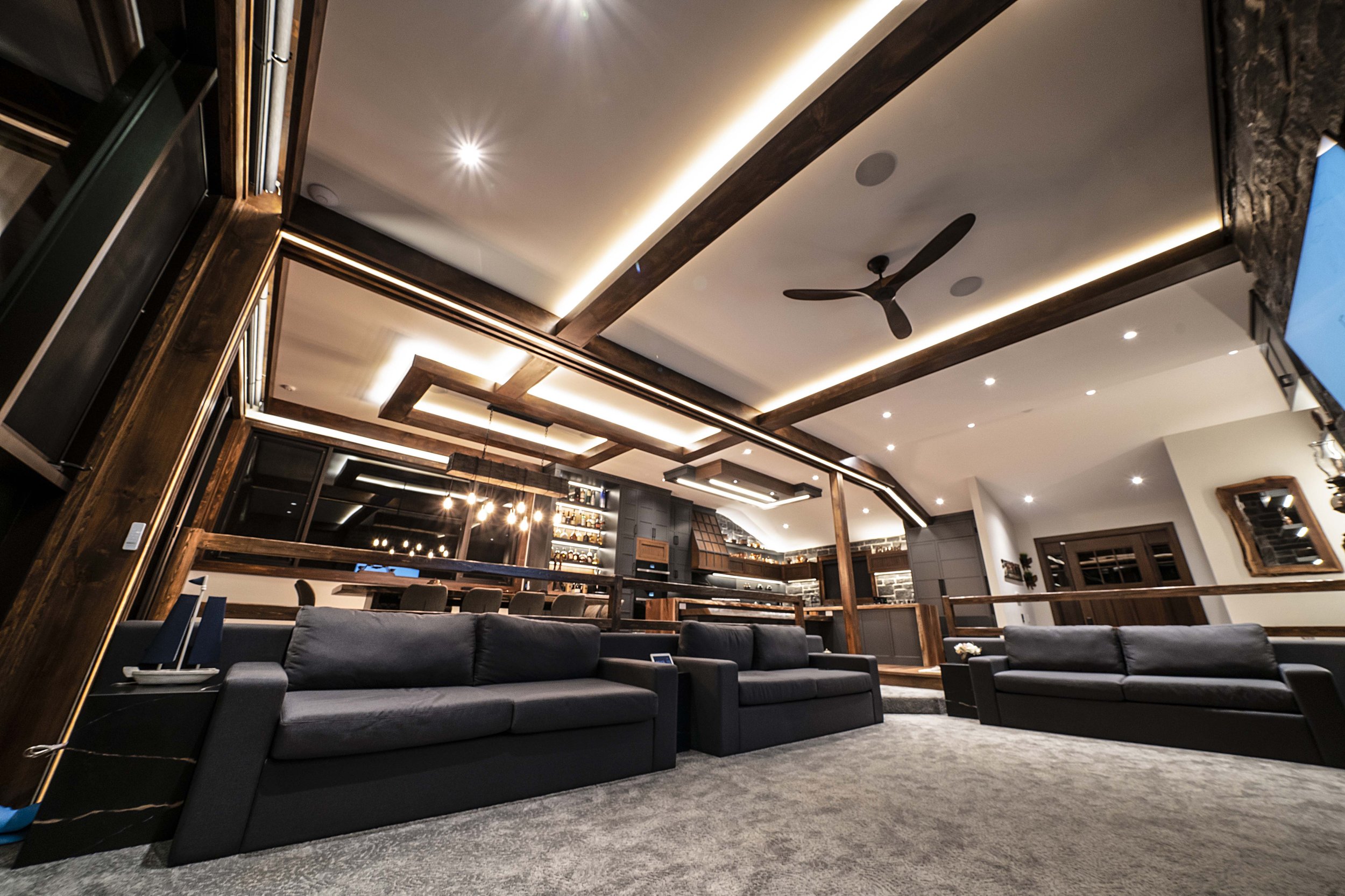 Modern living room with black sofas, large windows, and ceiling lights. Features a wood beam ceiling, fan, and adjacent kitchen area.