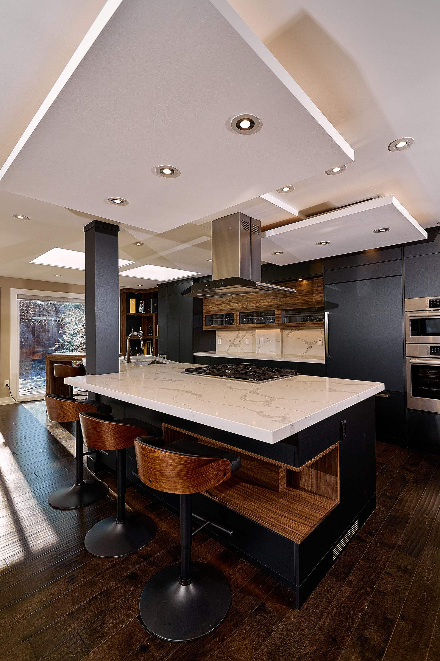 Modern kitchen with a large white marble island, sleek black cabinetry, stainless steel appliances, and wooden bar stools on dark hardwood flooring.