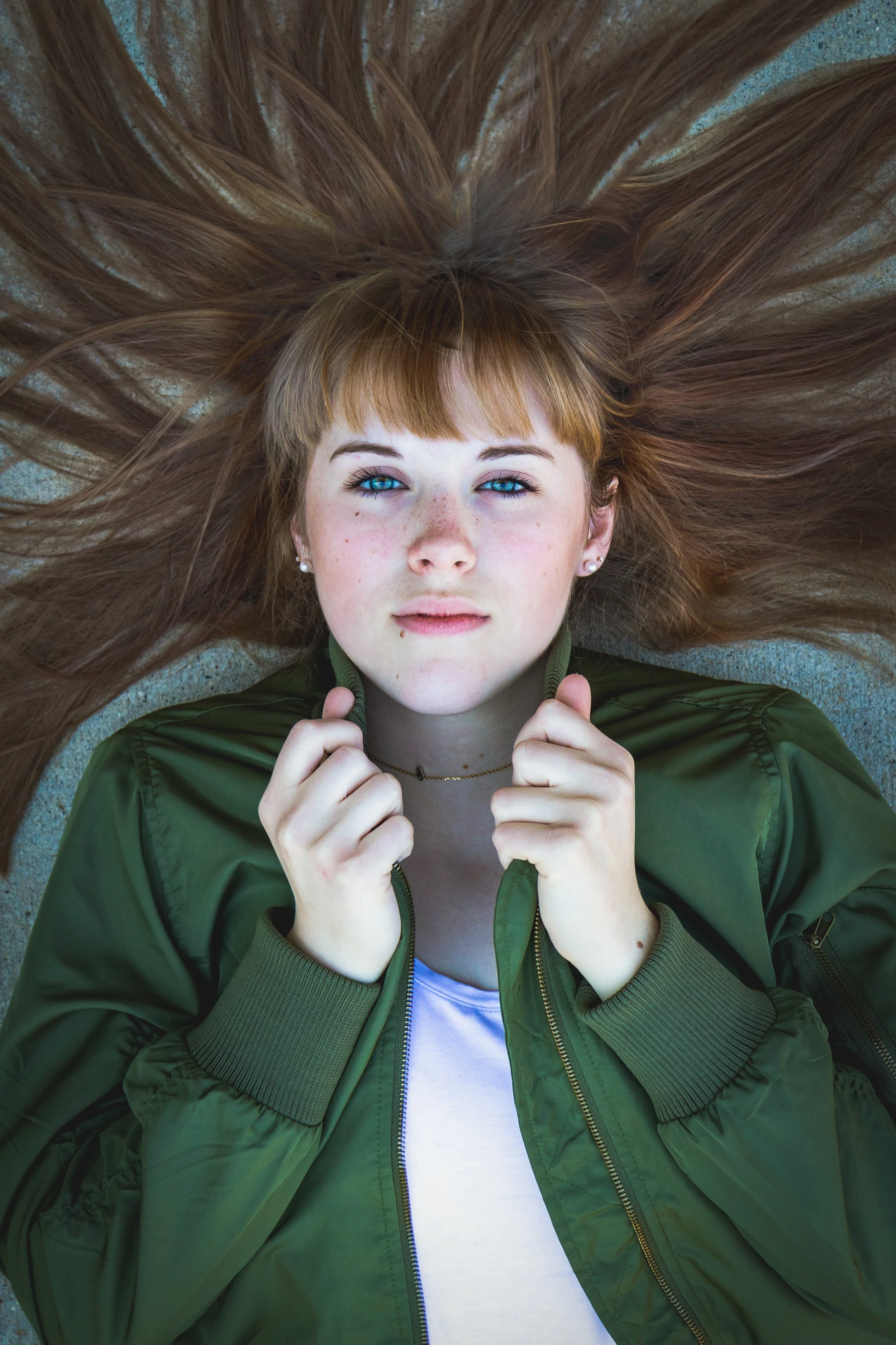 Young woman with long hair lying down, wearing a green jacket and a white shirt, looking at the camera.