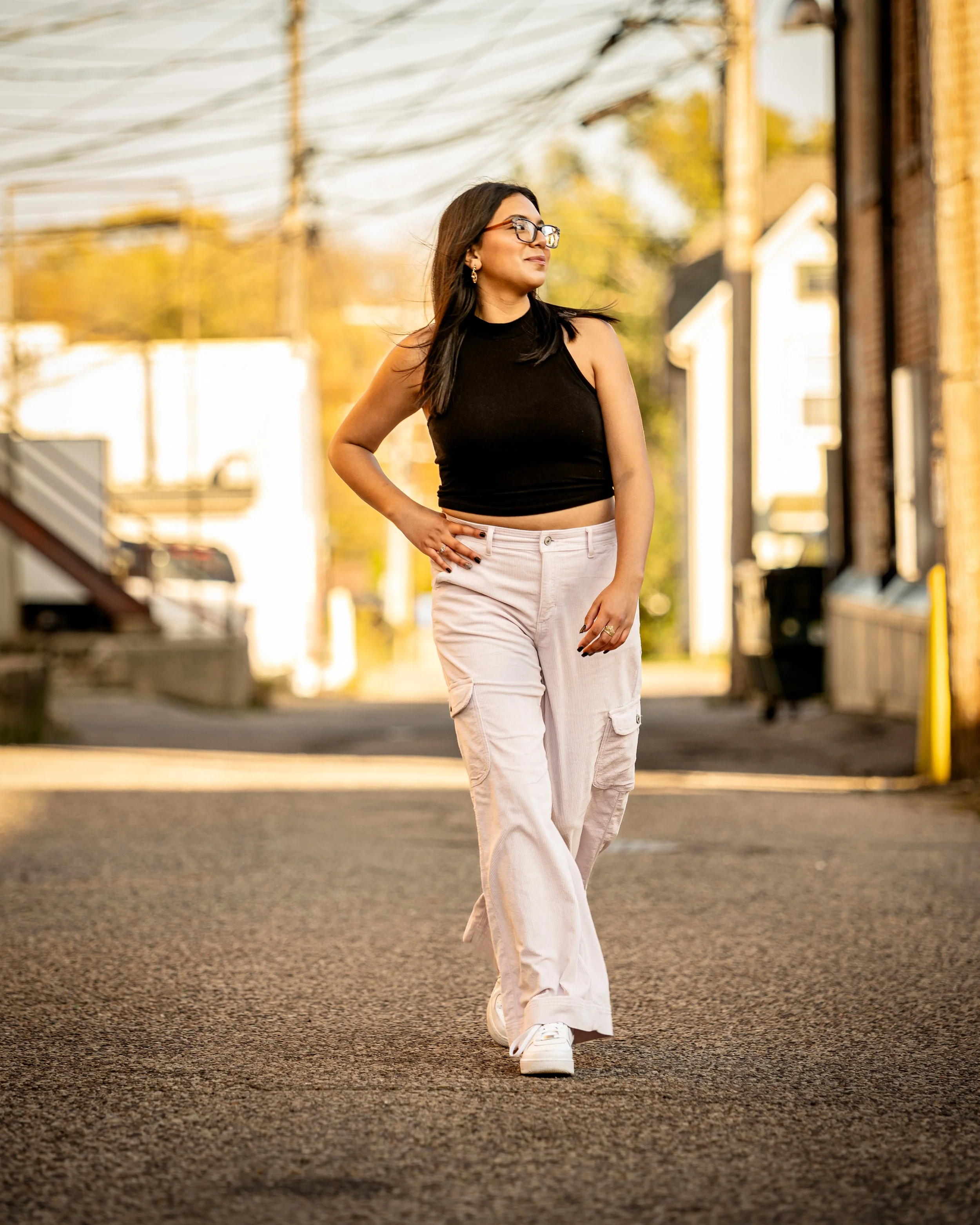 Woman walking confidently in an urban alleyway, wearing a black sleeveless top, white cargo pants, and glasses, with a relaxed expression.