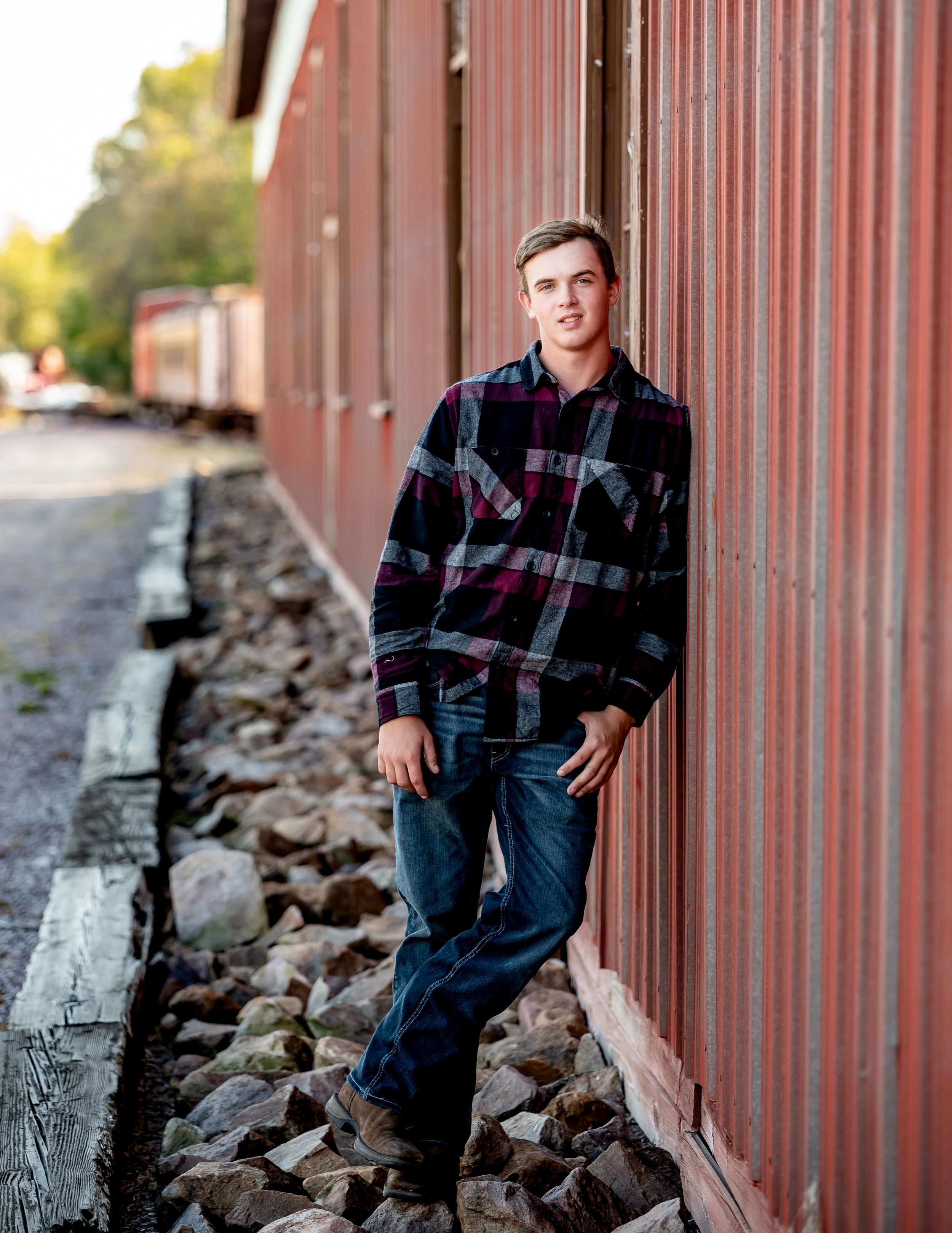 Young man in a checkered shirt leaning against a red corrugated wall, standing on rocky ground, with train tracks in the background.