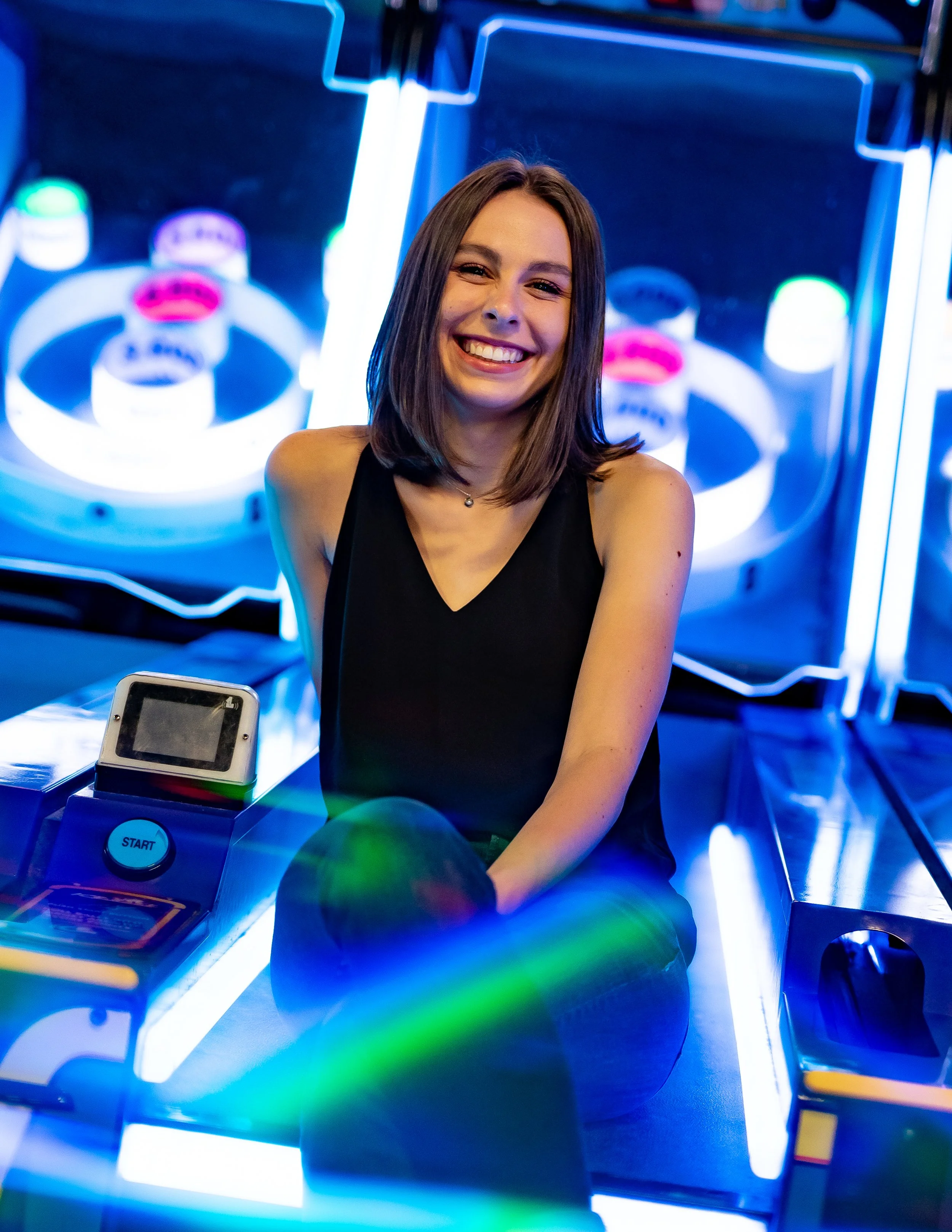 Woman smiling while sitting on a skee-ball machine in an arcade with colorful lights.