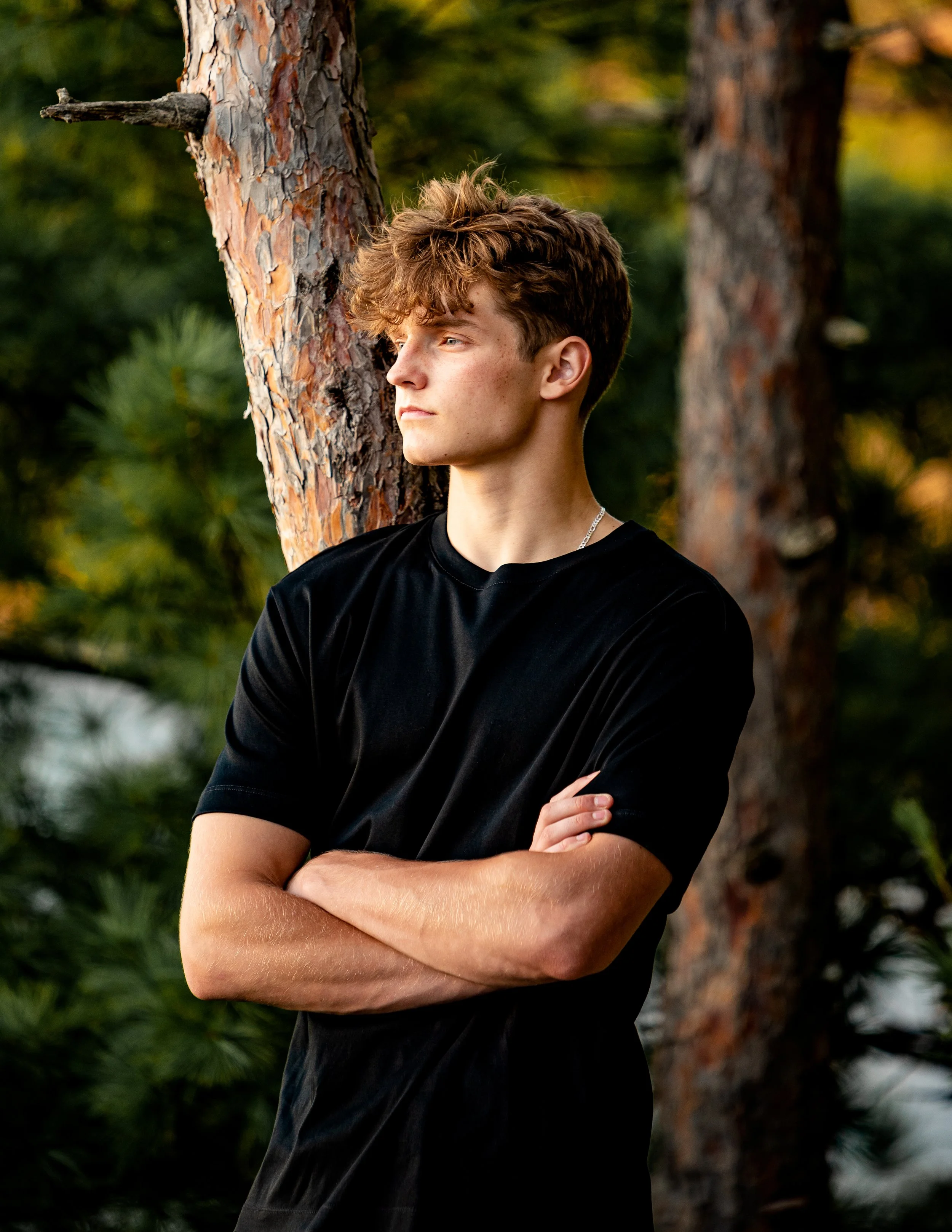 Young man in a black shirt standing with arms crossed, leaning against a tree in a forest setting during sunset for senior portraits near Baraboo, WI.