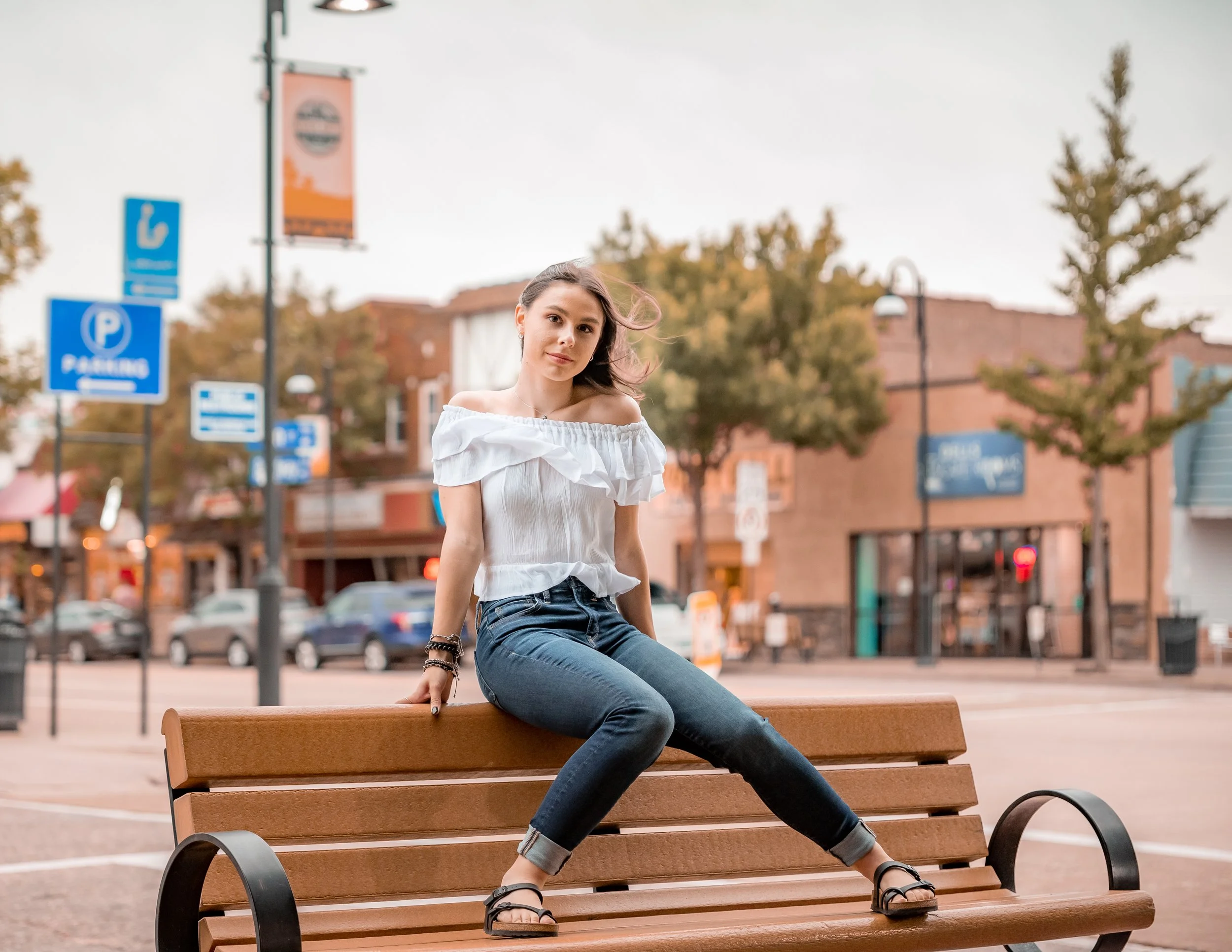 Young woman sitting on a bench in an urban setting, wearing a white off-shoulder shirt and jeans, with parking signs and buildings in the background.