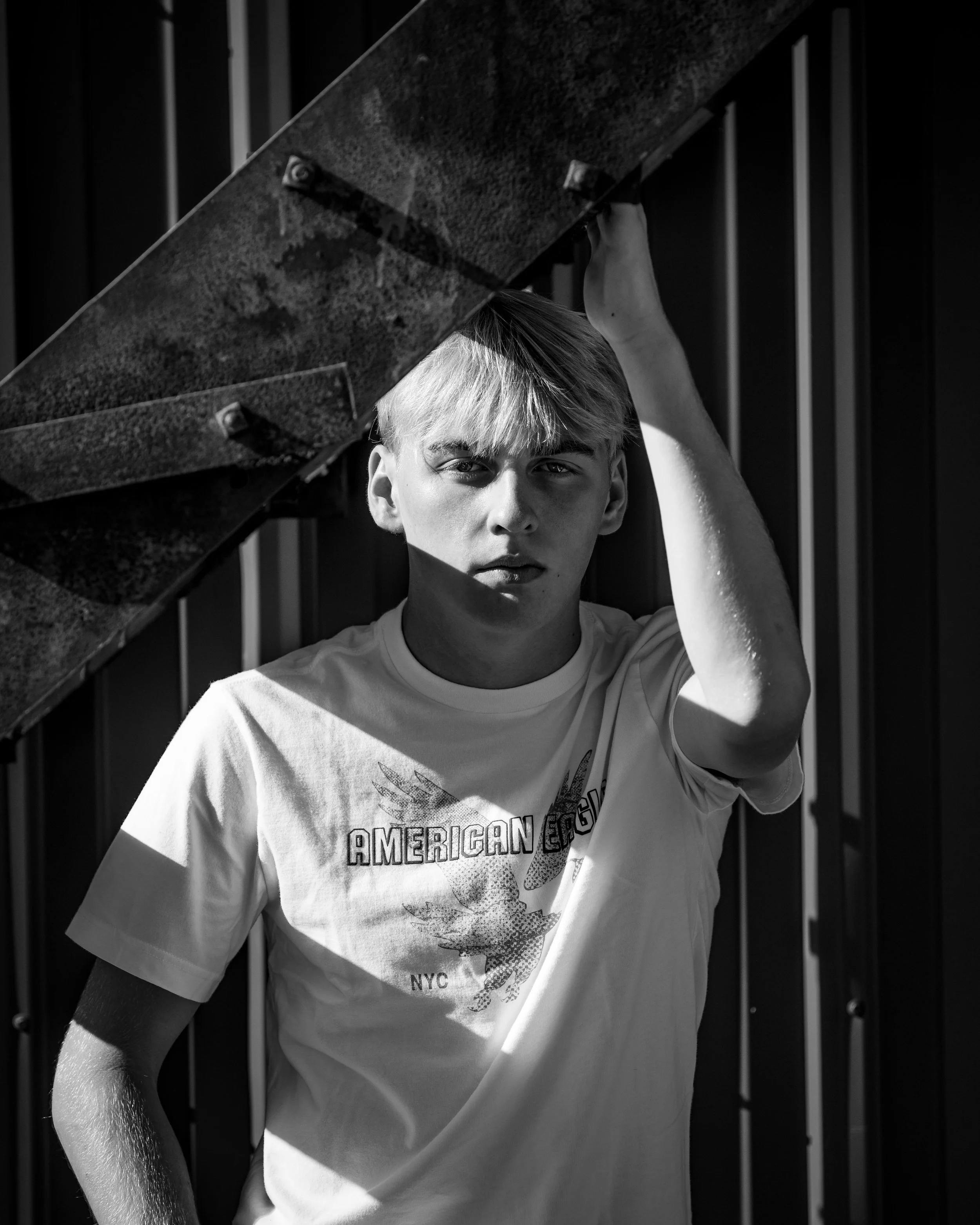 Black and white photo of a person with light-colored hair, wearing a T-shirt, standing outdoors by a metal structure.