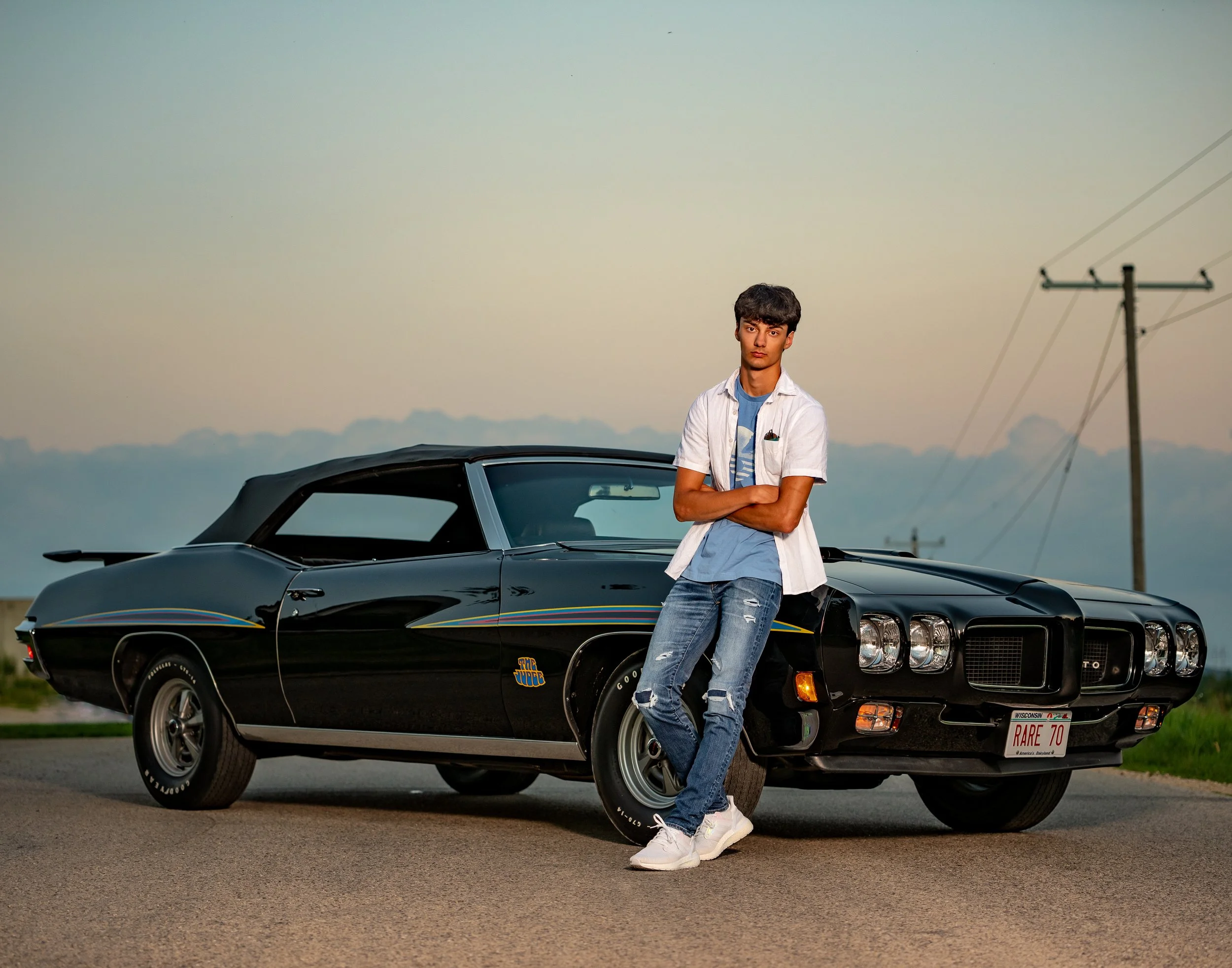 A person leaning against a black classic car on a road during sunset.