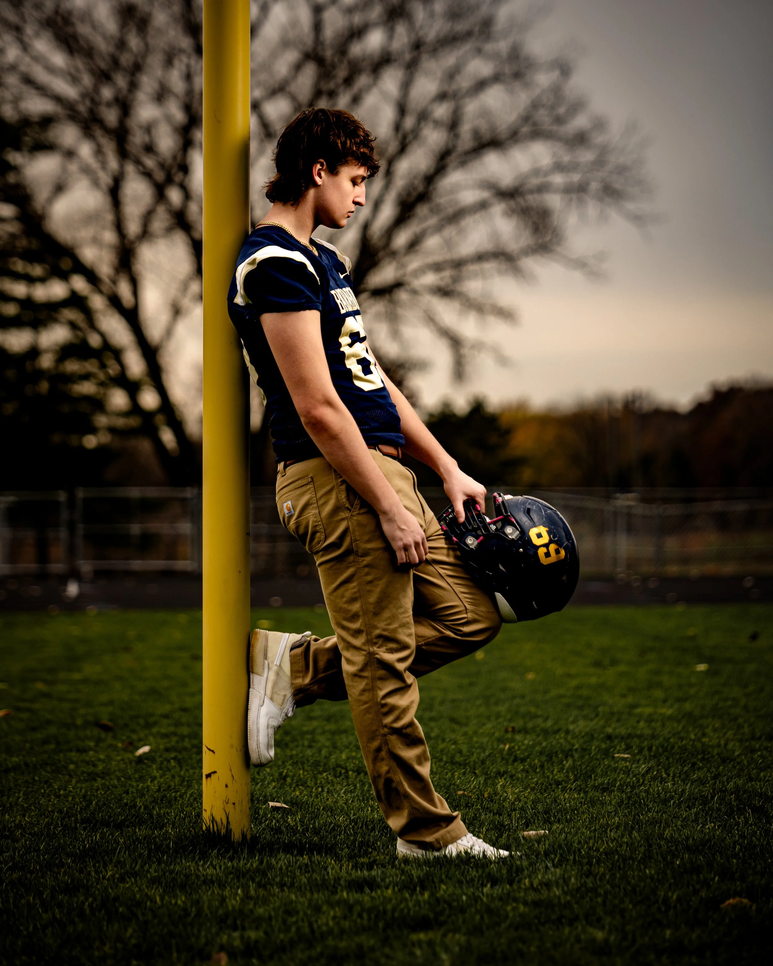A young man in a blue football jersey and khaki pants leaning against a yellow goal post on a grassy field, holding a football helmet, with trees in the background.