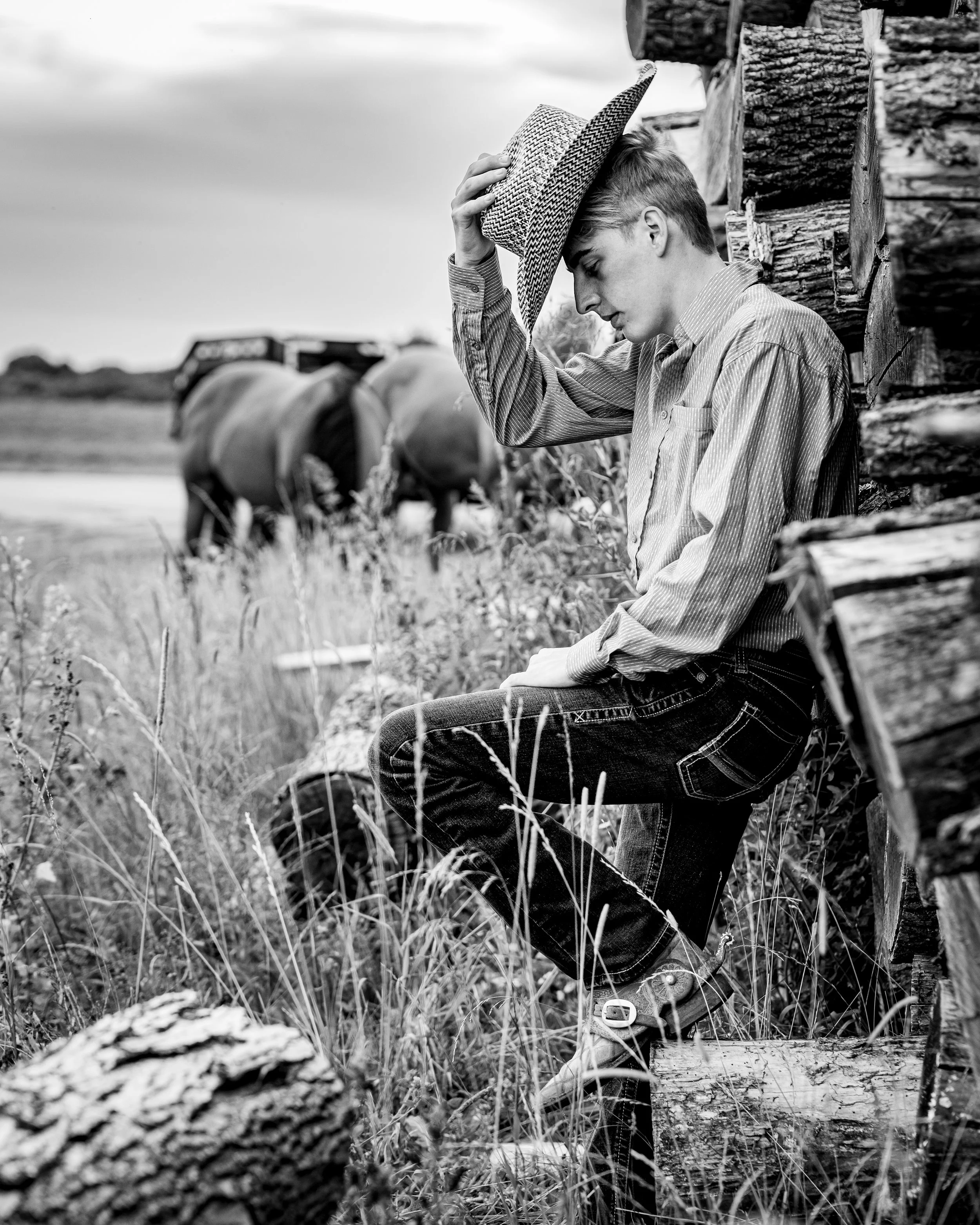 Black and white image of a person in a cowboy hat sitting on a stack of logs in a field, with horses grazing in the background.