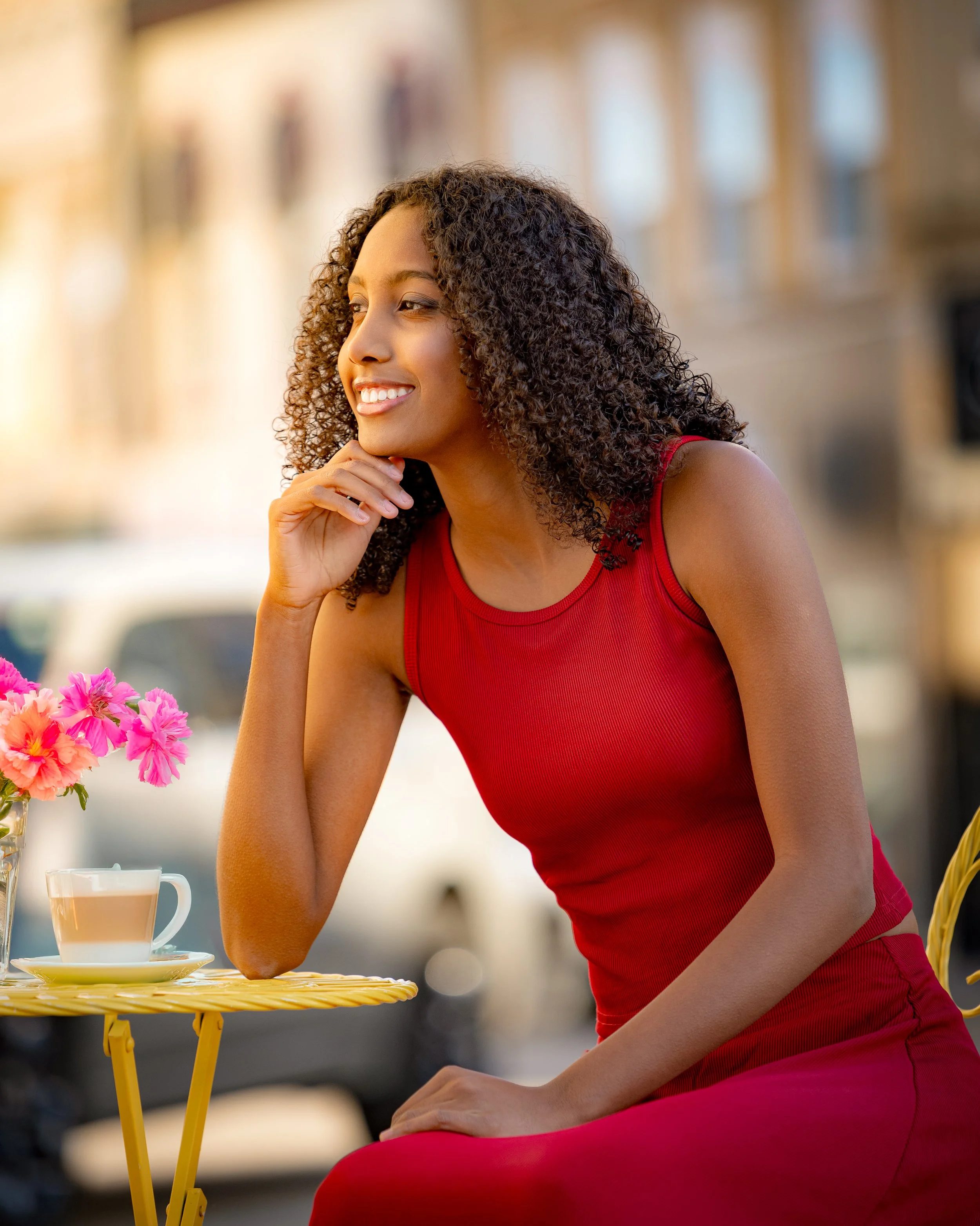 A young woman in a red dress sitting at an outdoor cafe table with a cup of tea and pink flowers for her cafe themed senior pictures in Baraboo, WI.