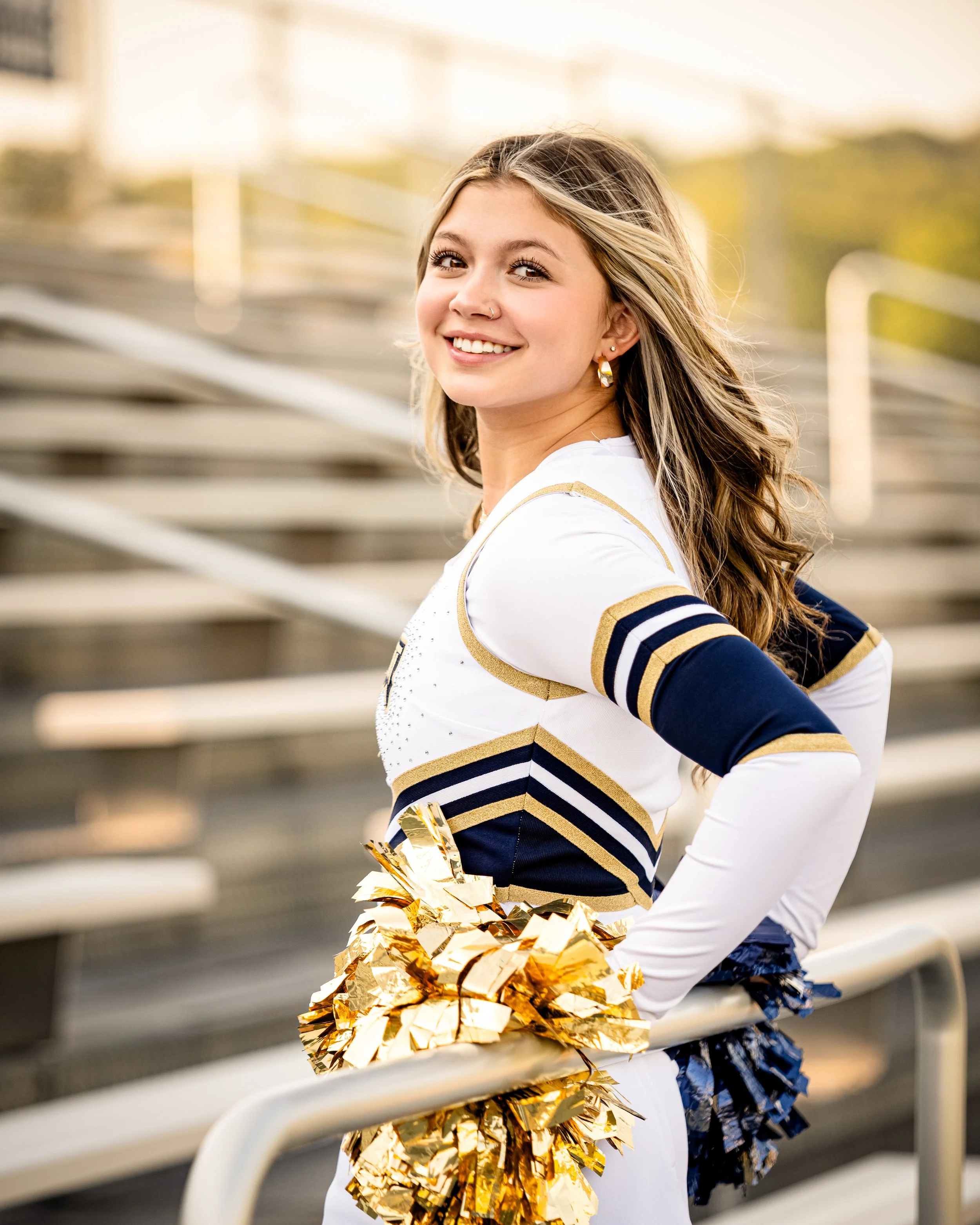 Baraboo Cheerleader in white and blue uniform smiling with golden pom-poms in bleachers at sunset for her senior photos.