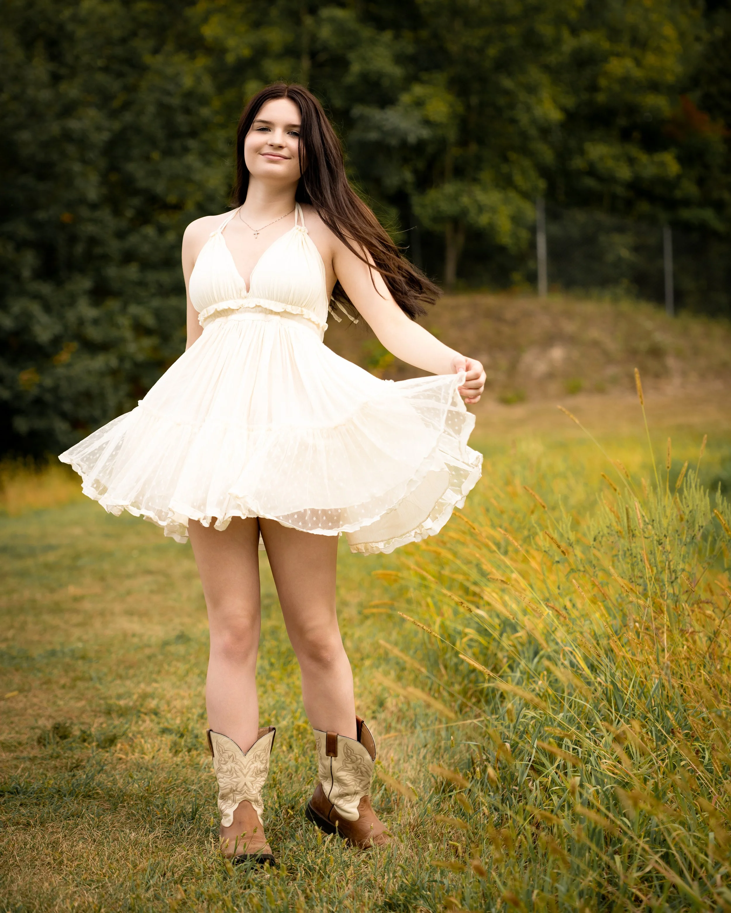 Young woman in white dress and cowboy boots standing on grass in a field, with green trees in the background.