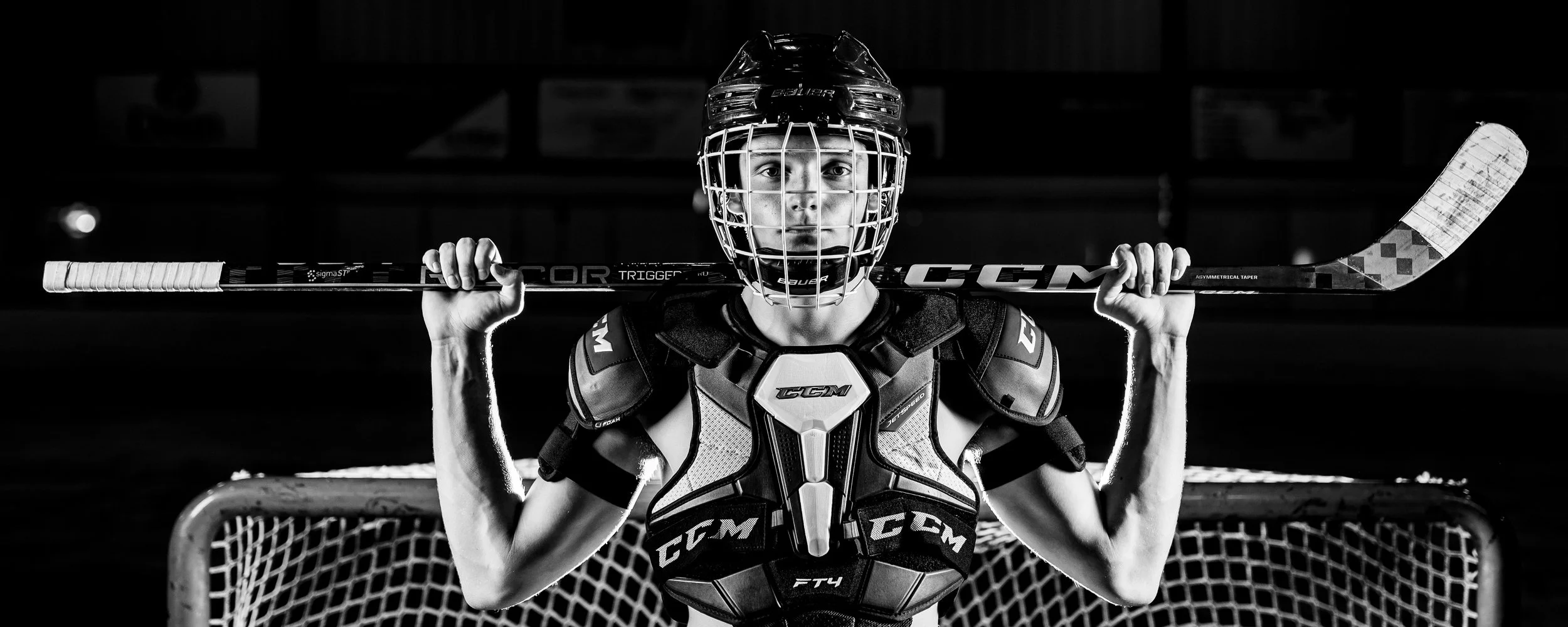 Black and white image of a hockey player holding a stick across their shoulders, wearing protective gear and standing in front of a goal net.