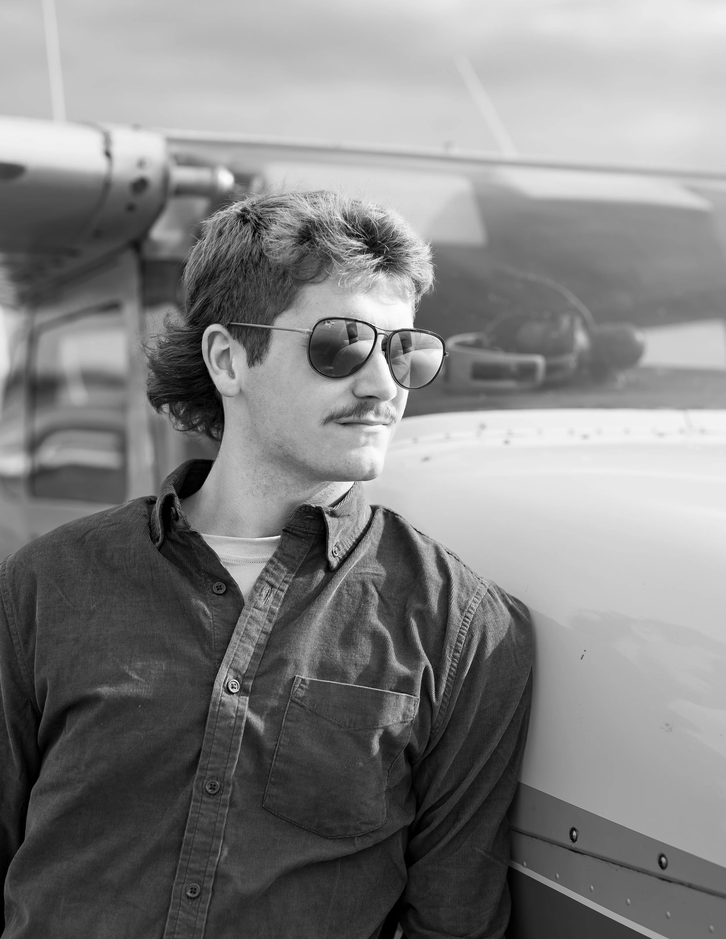 Black and white photo of a man with sunglasses and a mustache, leaning against a small airplane.
