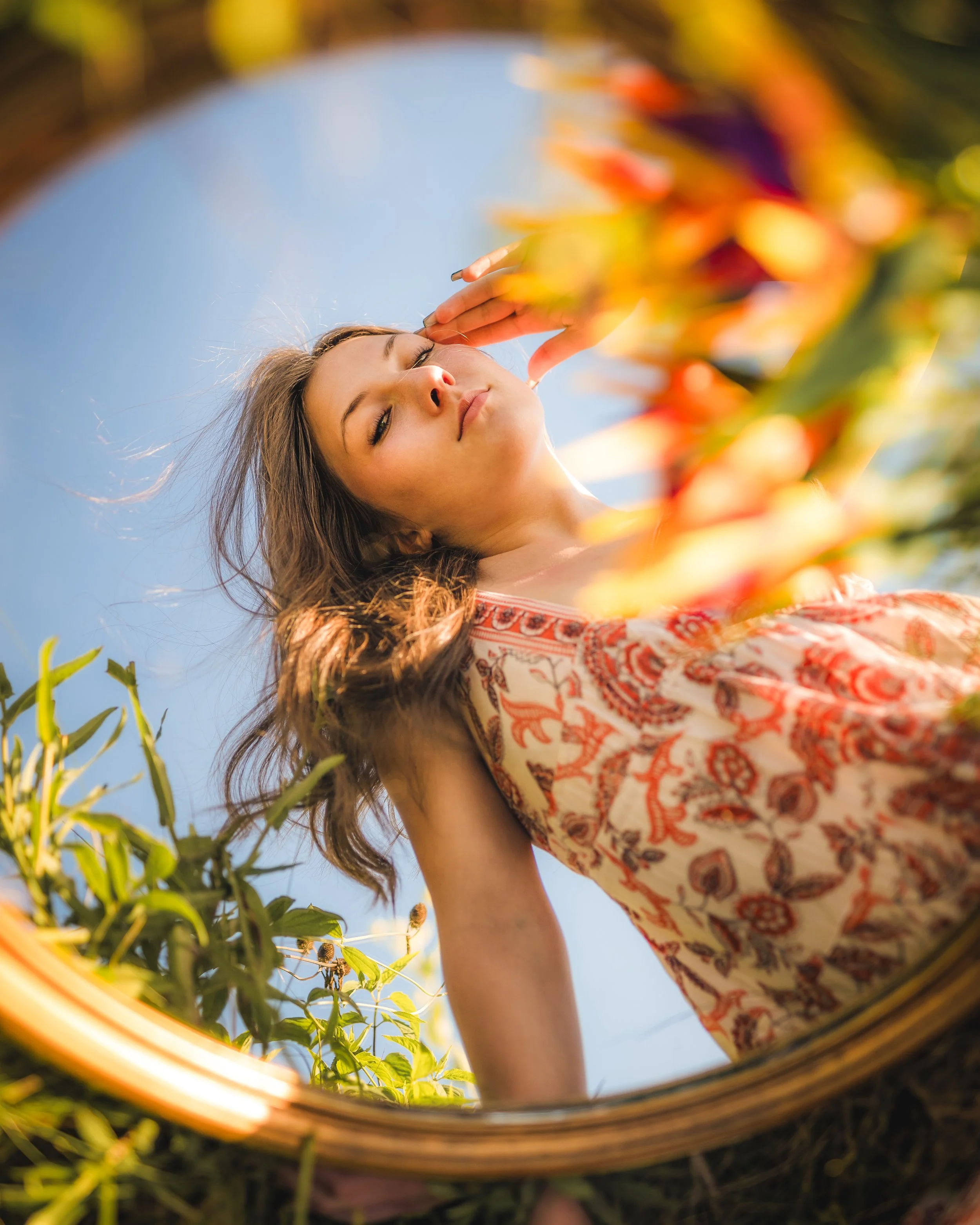 Woman looking at her reflection in a round mirror outdoors, surrounded by colorful plants, wearing a patterned dress for her floral themed senior pictures near Baraboo, WI.